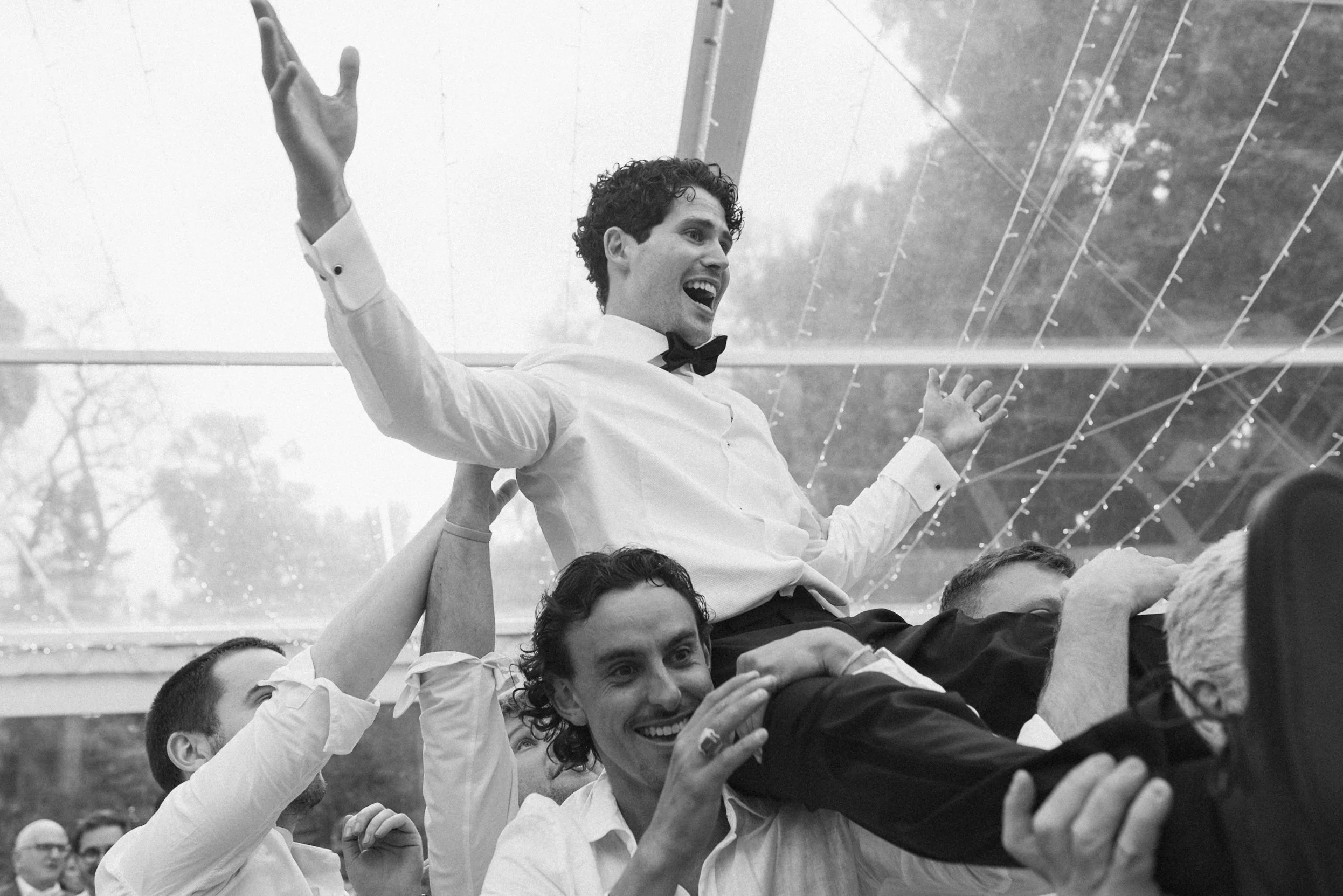 A group of people celebrating outdoors, with a man in a tuxedo and bow tie sitting on someone's shoulders, raising his arms and smiling. The scene is lively and joyful, with other smiling individuals around him.