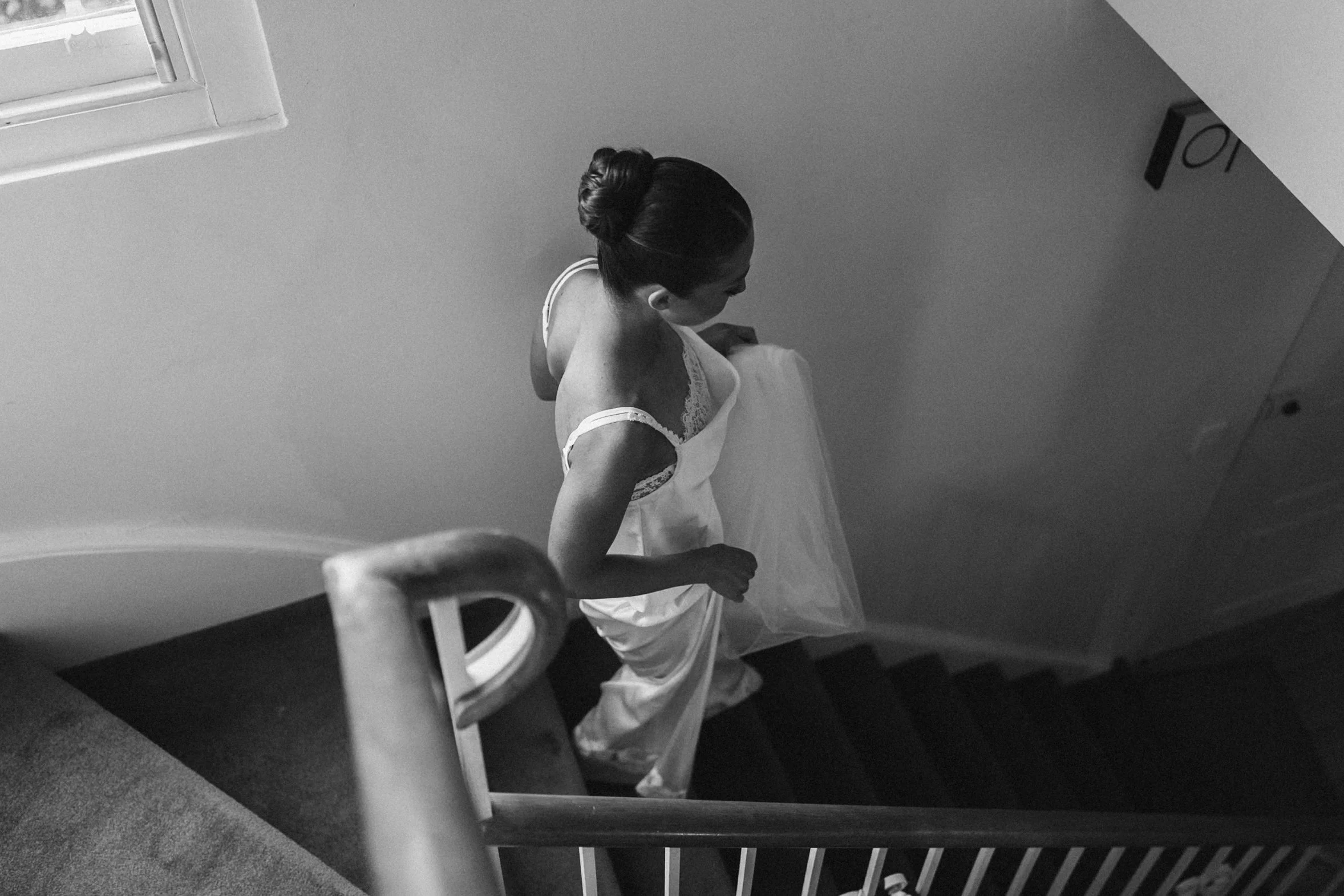 A woman with dark hair styled in an elegant bun, wearing a light-colored dress with lace details, stands in a room with white walls and a window, leaning slightly to her right, and holding a piece of cloth, possibly a dress, near her waist.