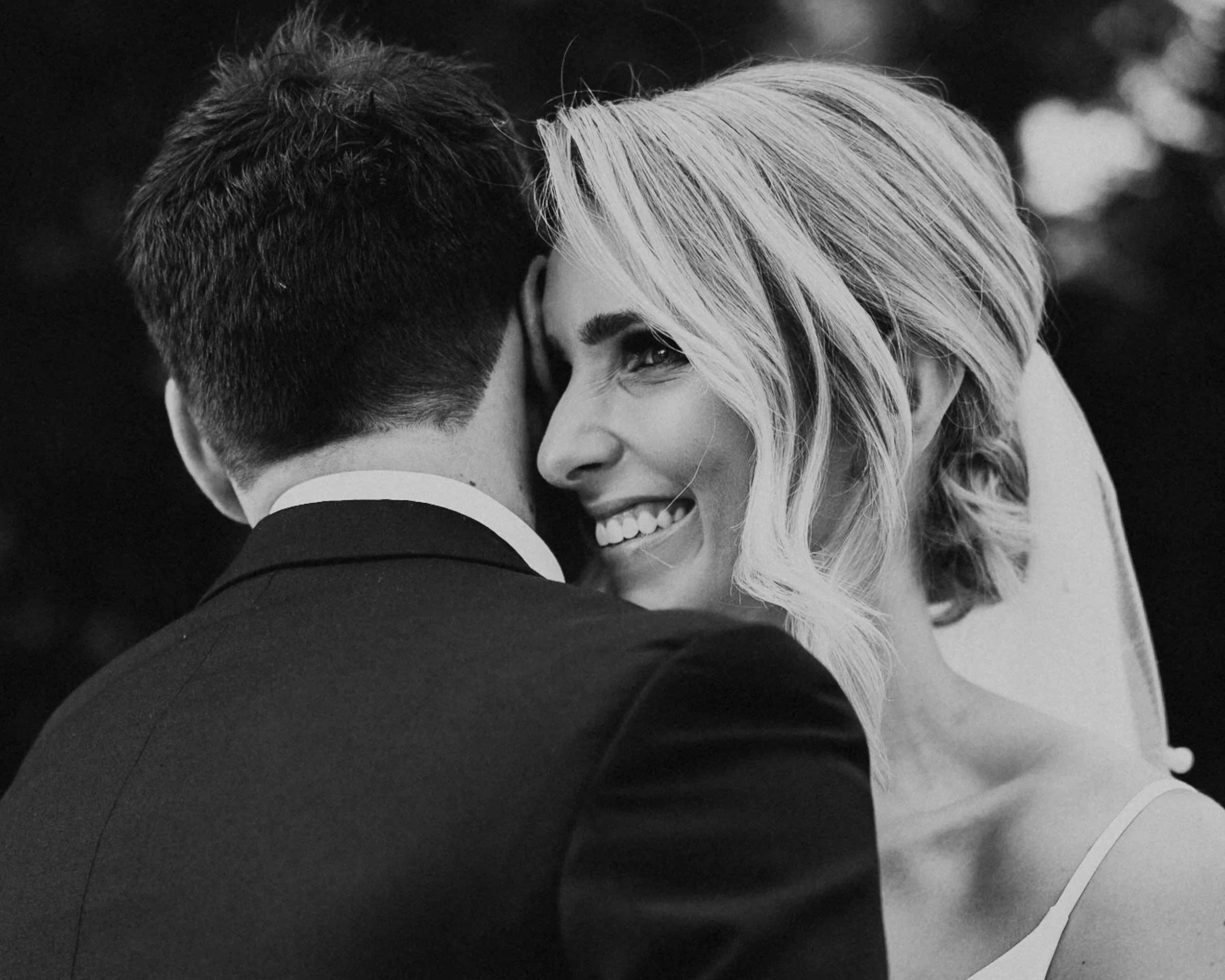 Black and white photo of a man and woman embracing closely, with the man’s back to the camera and the woman smiling and looking sideways.