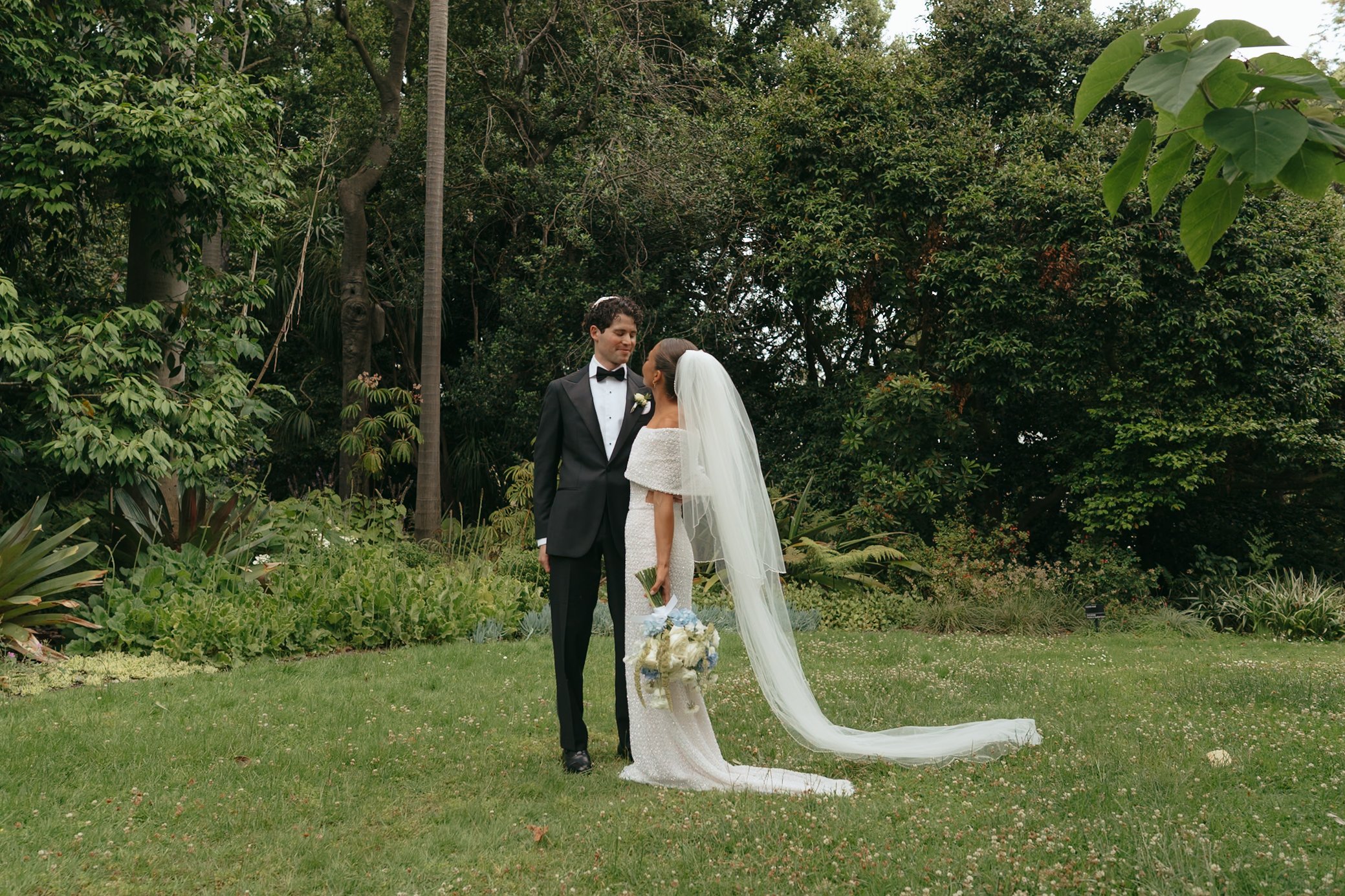 A bride and groom standing in a garden, gazing at each other during their wedding ceremony. The groom wears a black tuxedo with a bow tie, and the bride wears a white wedding gown with a veil. The bride is holding a bouquet of flowers, and behind the