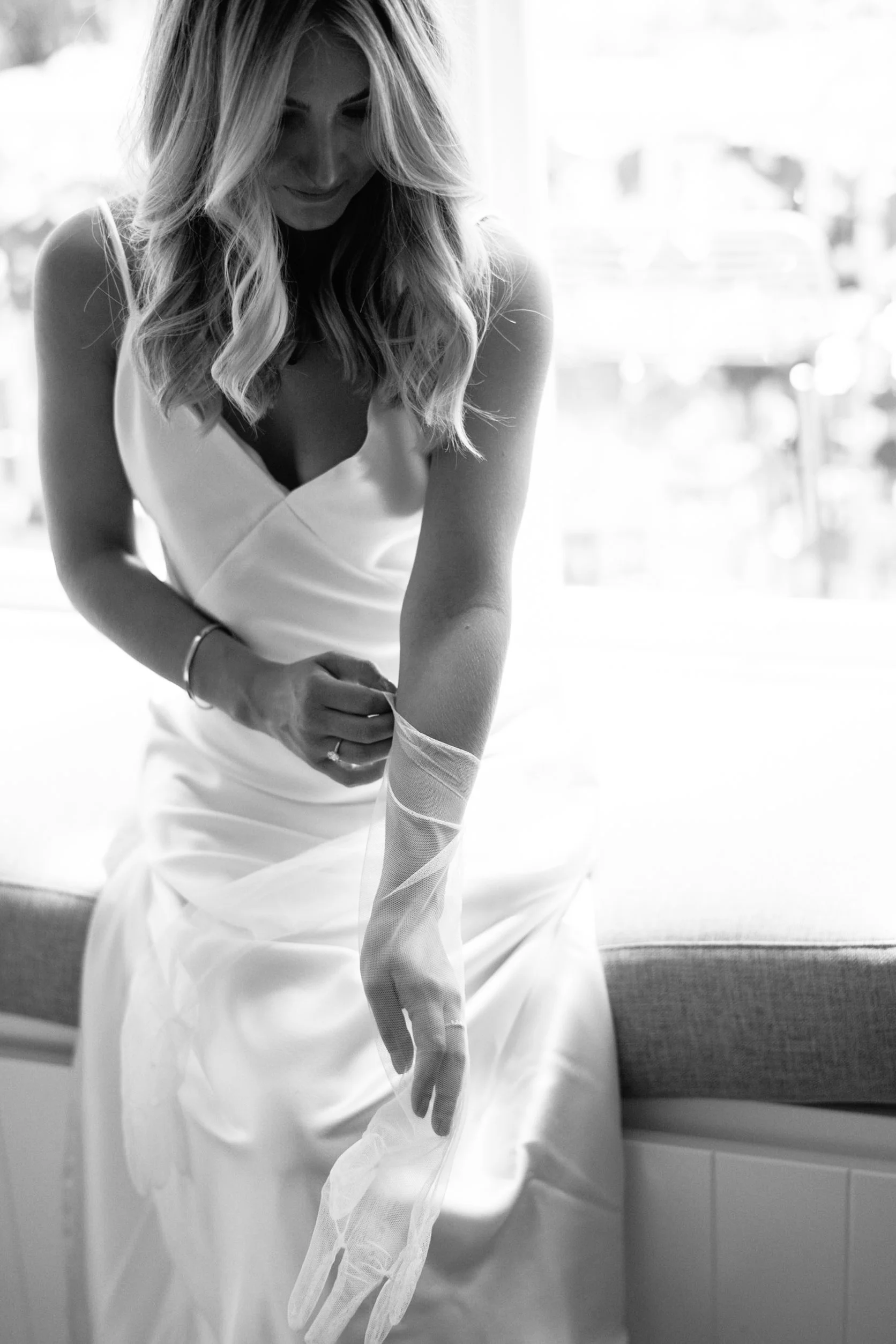 Black and white photo of a woman in a white dress putting on a long white glove, sitting on a bench or chair in a bright room.