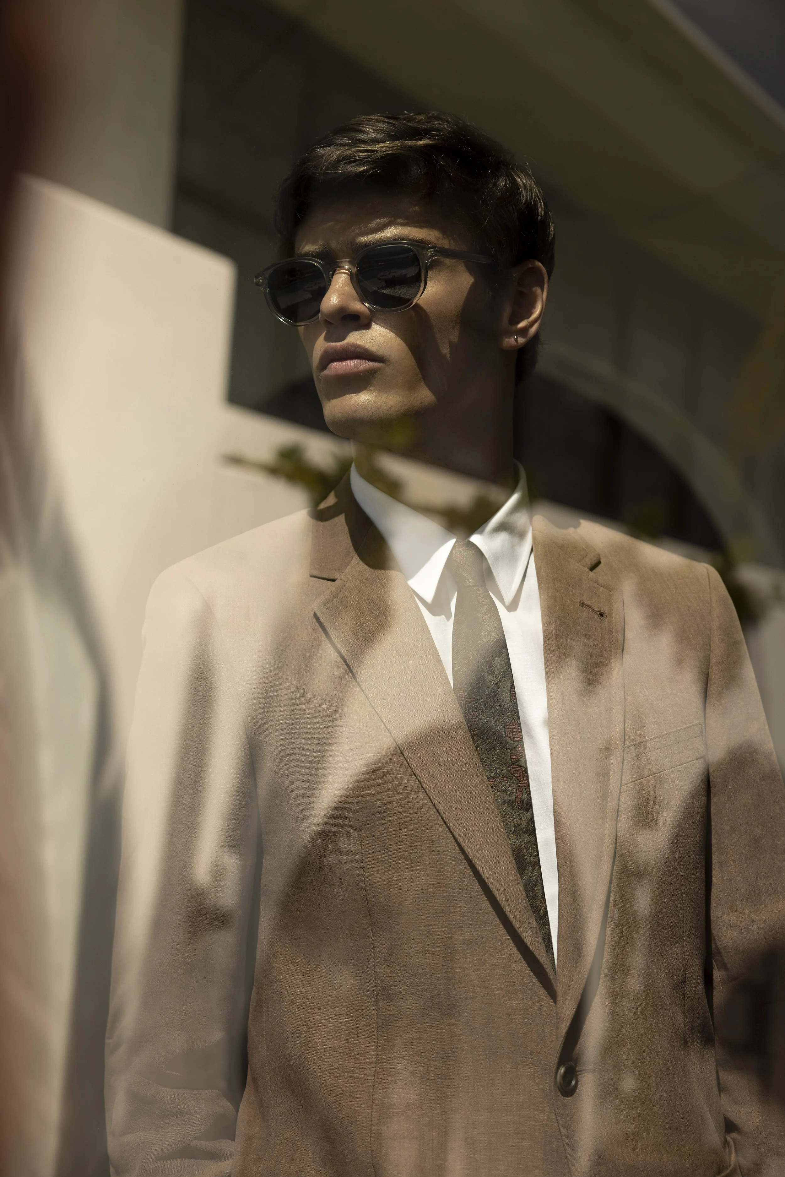 A man wearing dark sunglasses and a tan blazer looks away as reflected in a glass window.