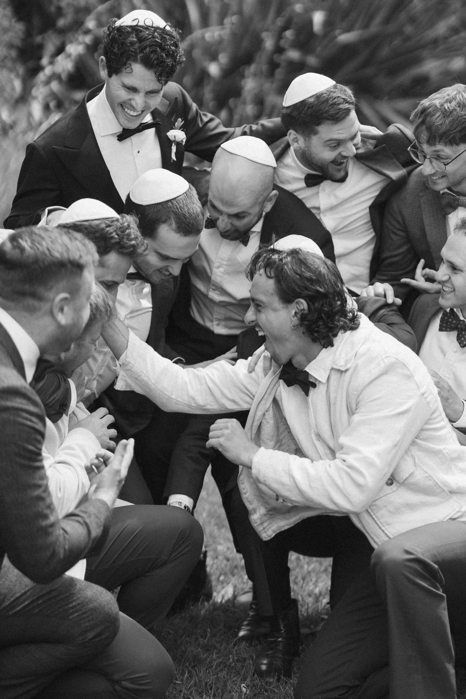 Black and white photo of men in tuxedos and dresses, wearing yarmulkes, joyfully celebrating outdoors with some men kneeling or sitting on grass, others standing around, smiling, embracing, and raising their arms in happiness.