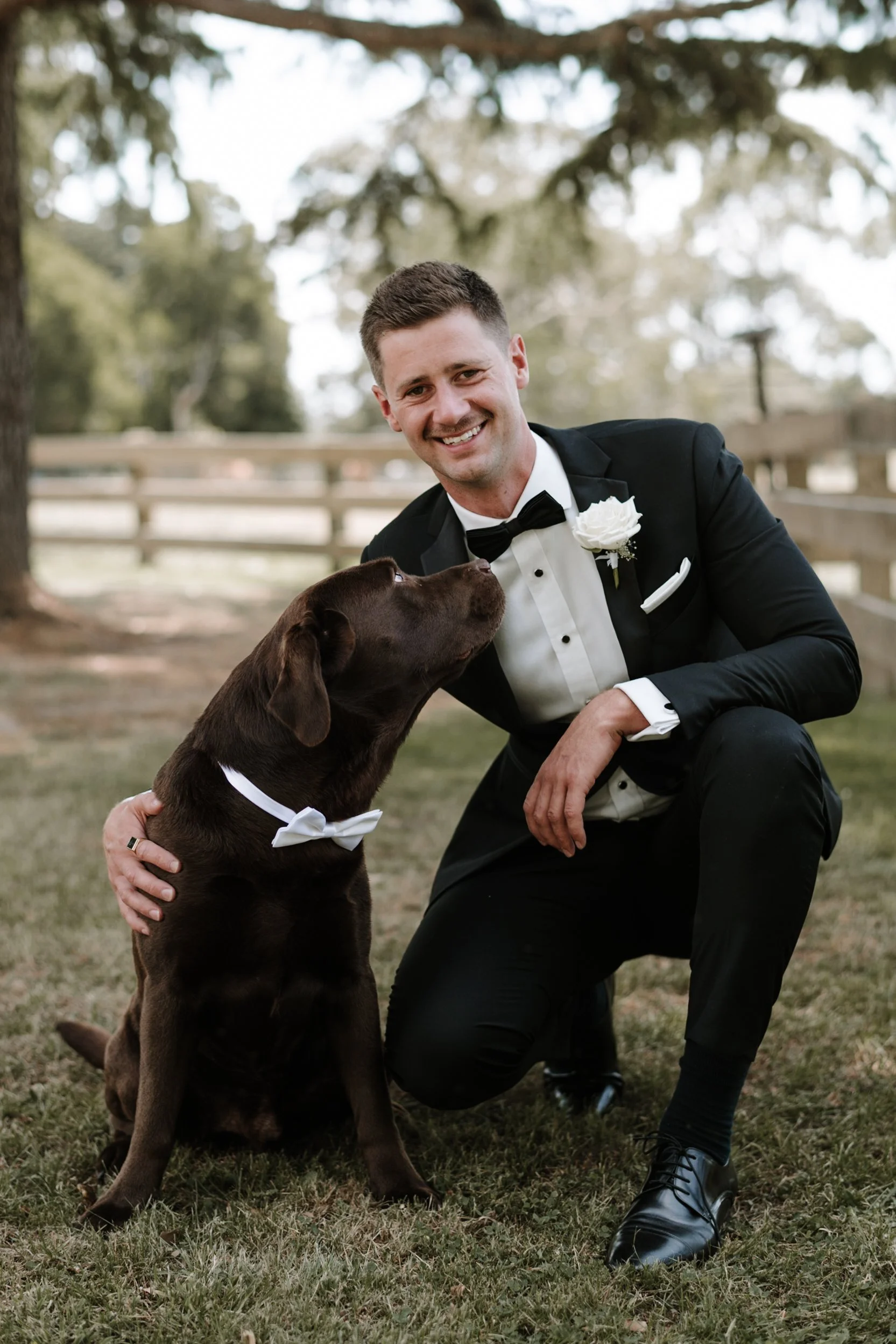 A man in a tuxedo and bow tie kneeling outdoors, smiling at a brown dog wearing a white bow tie, in a park-like setting with trees and a wooden fence in the background.
