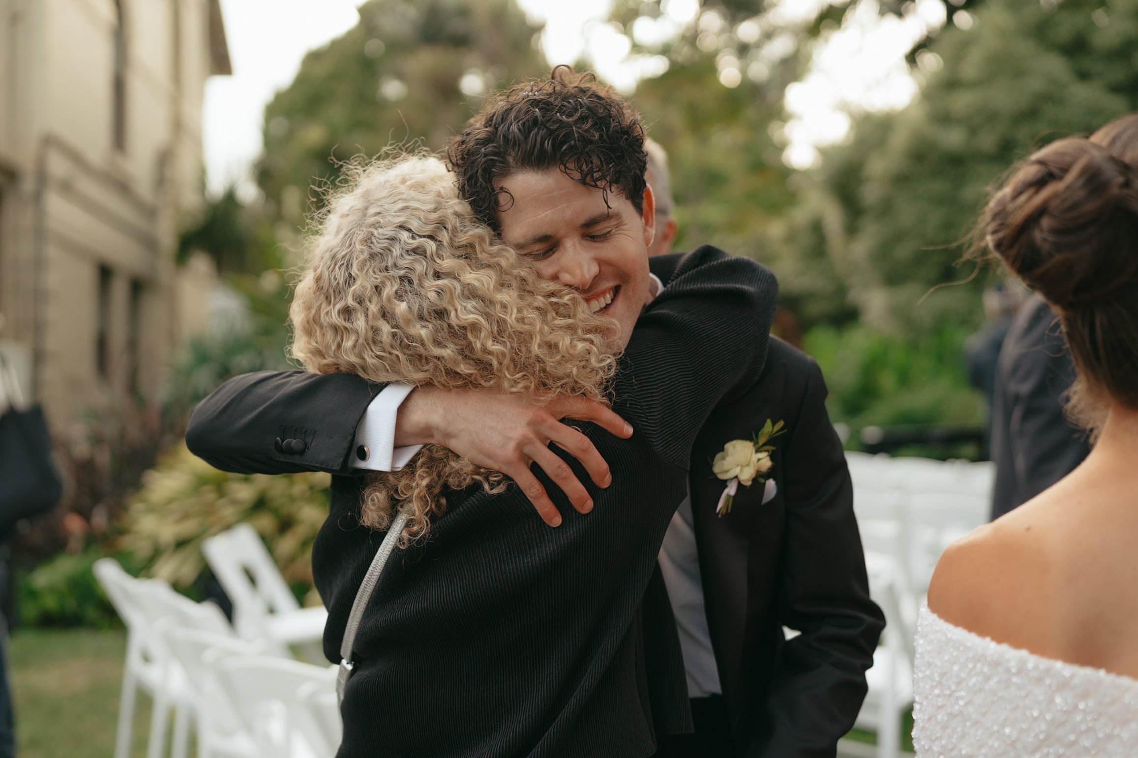 A man with curly dark hair hugging a woman with blonde curly hair at an outdoor wedding ceremony, with white chairs and trees in the background.