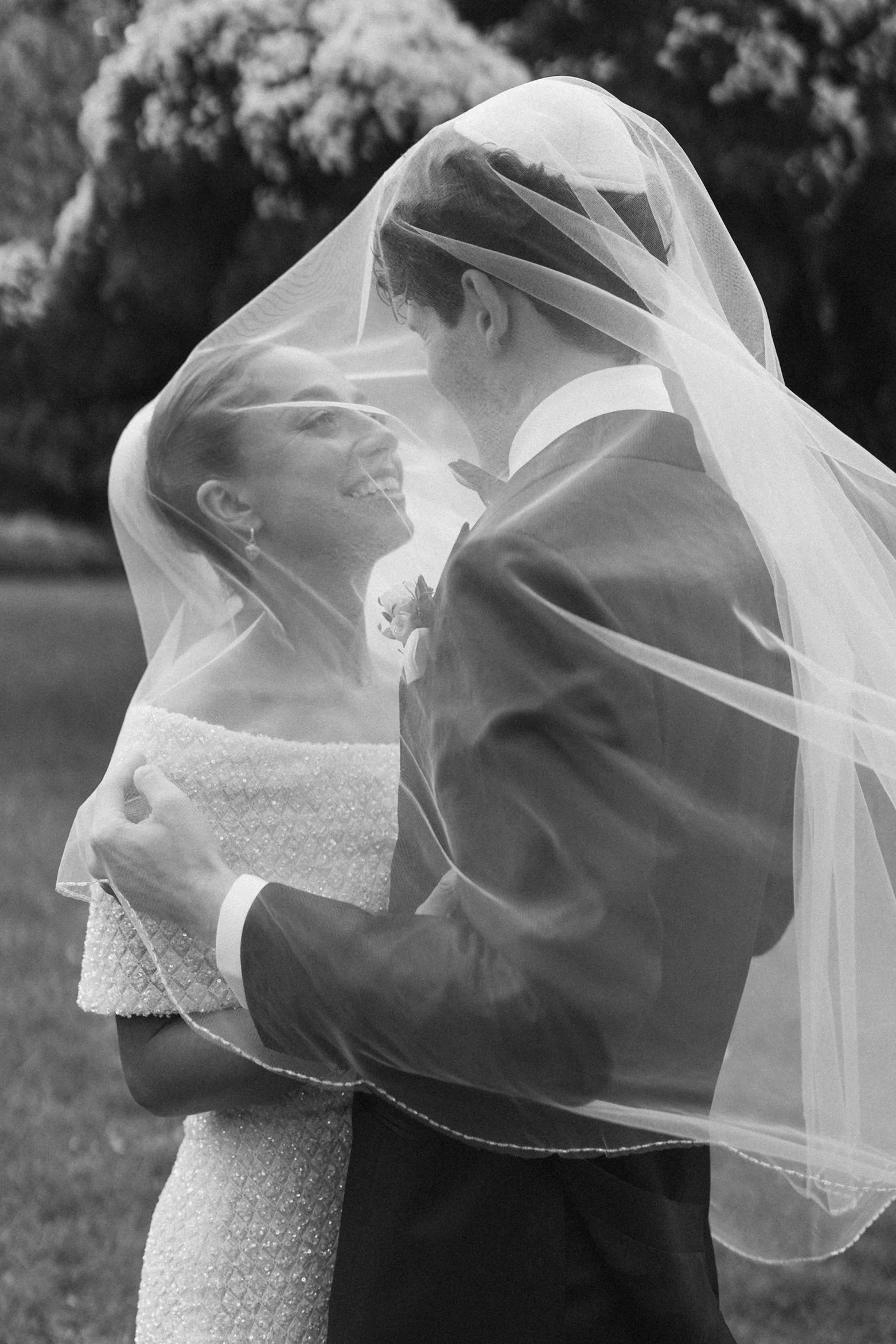 A black-and-white photo of a couple in wedding attire, sharing a joyful moment under a wedding veil, with the bride smiling at the groom outdoors.