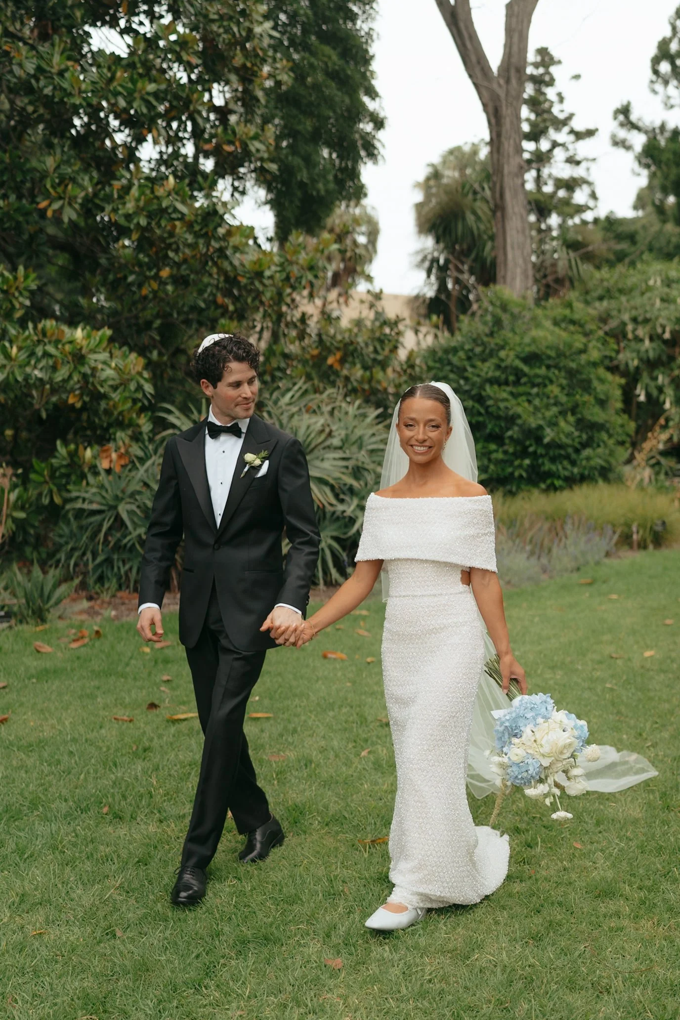 A bride and groom walking hand in hand through a lush garden during their wedding. The bride is wearing a white off-shoulder wedding gown and holding a bouquet of white and light blue flowers. The groom is dressed in a black tuxedo with a bow tie and
