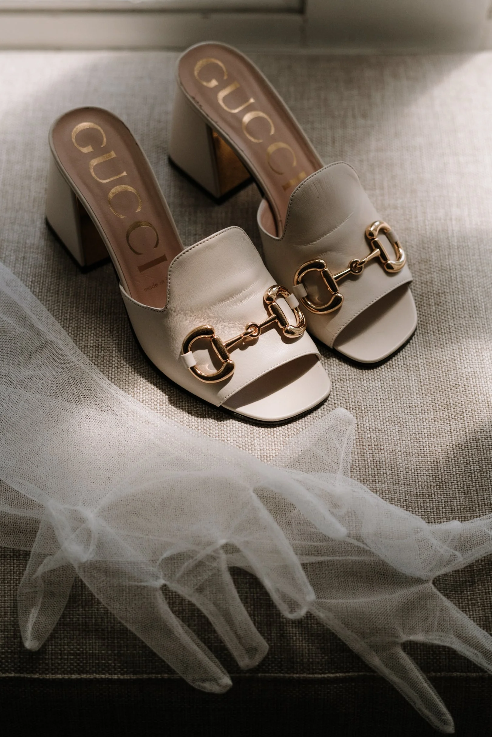 A pair of white Gucci high-heeled sandals with gold hardware, placed on a textured fabric surface with tulle fabric in the foreground.