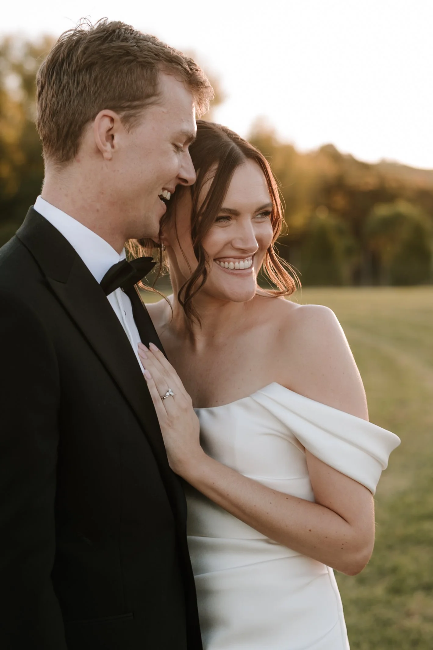 A newlywed couple standing close together outdoors during sunset, smiling happily. The groom is in a black tuxedo with a bow tie, and the bride is in a white off-the-shoulder wedding dress with wavy brown hair.