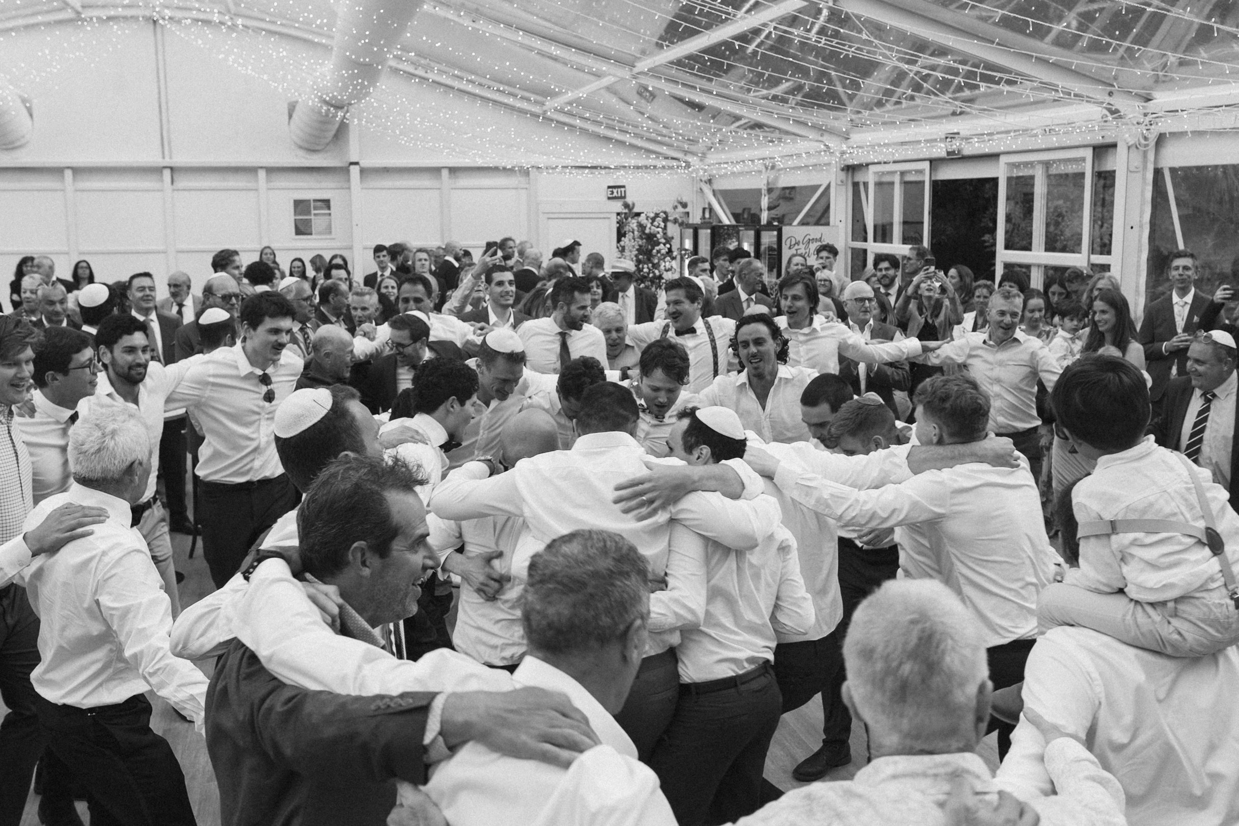 A group of men dressed in white shirts and dark pants are participating in a traditional Jewish celebration or dance indoors under string lights, with some wearing kippahs and others embracing or holding hands.