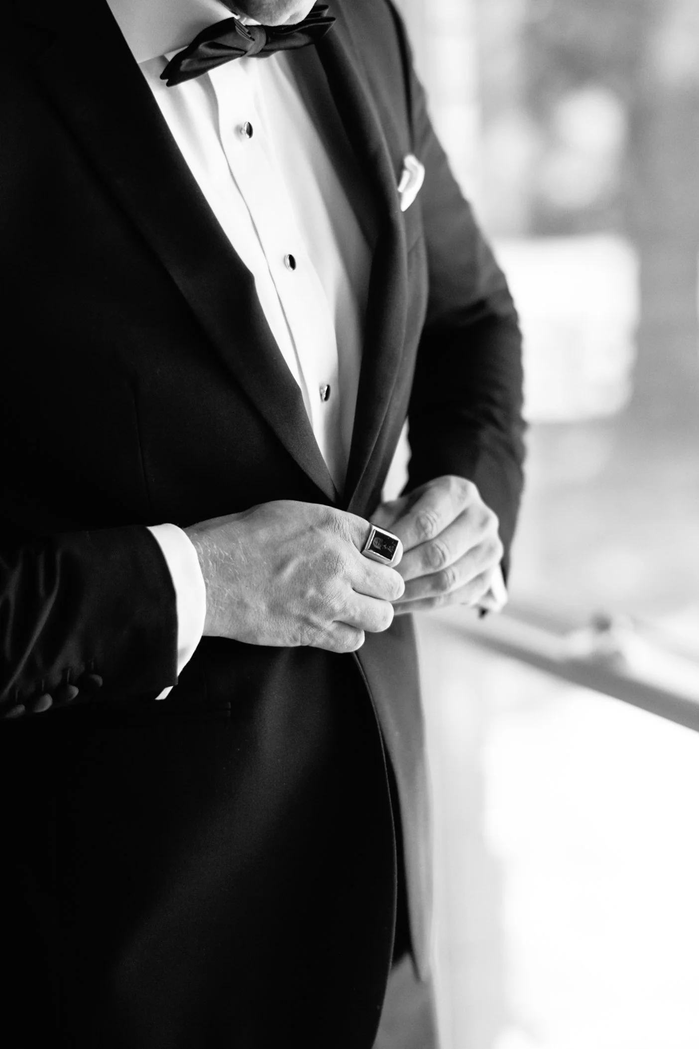 Close-up black and white photo of a man dressed in a tuxedo, with a bow tie, adjusting his cufflink.