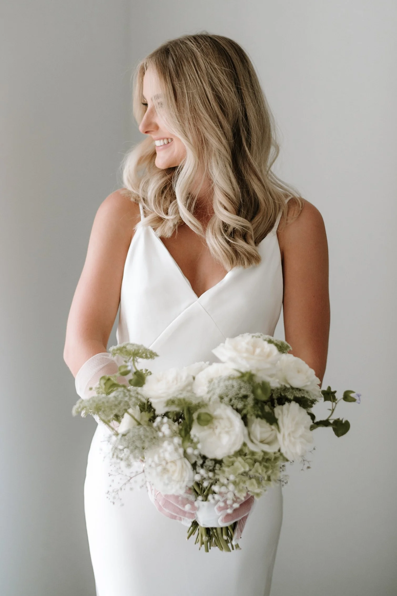 A woman with blonde wavy hair smiling and holding a large bouquet of white flowers, wearing a white sleeveless dress against a plain light gray background.