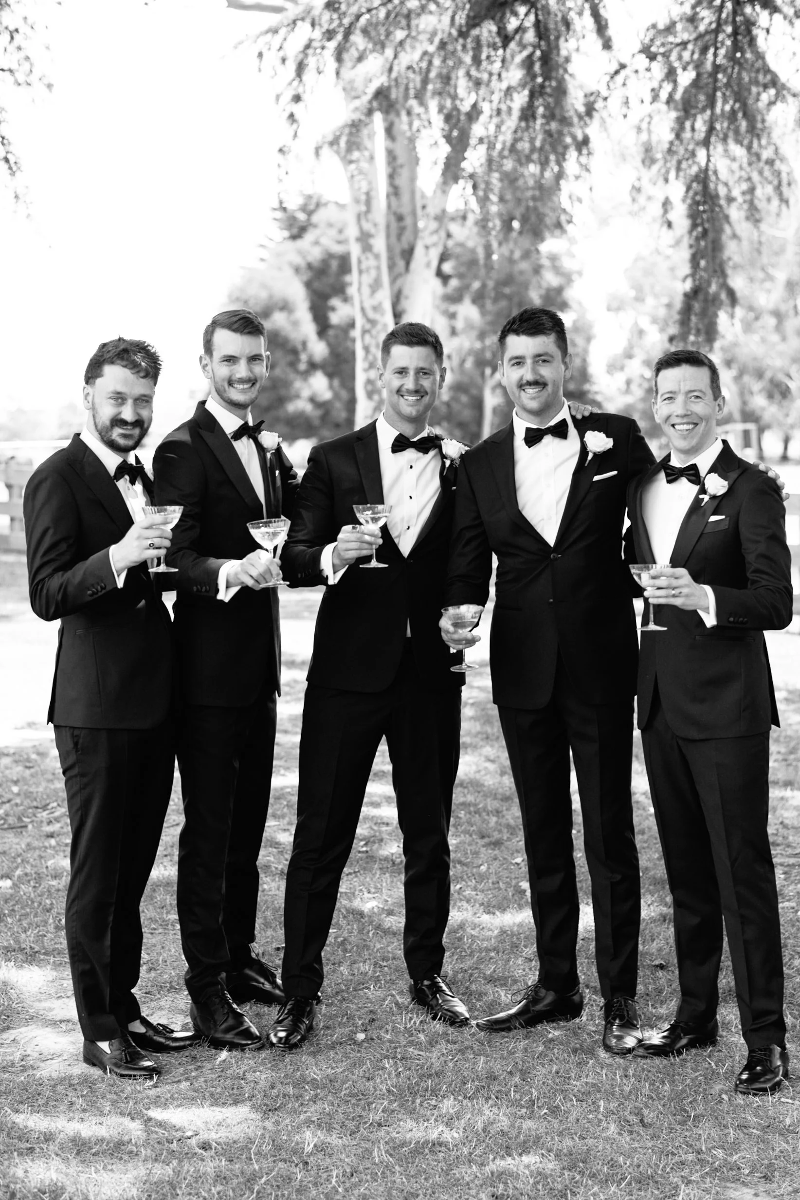 A group of six men in tuxedos with bow ties and boutonnieres, holding champagne glasses, standing outdoors on grass with trees in the background, smiling at the camera.
