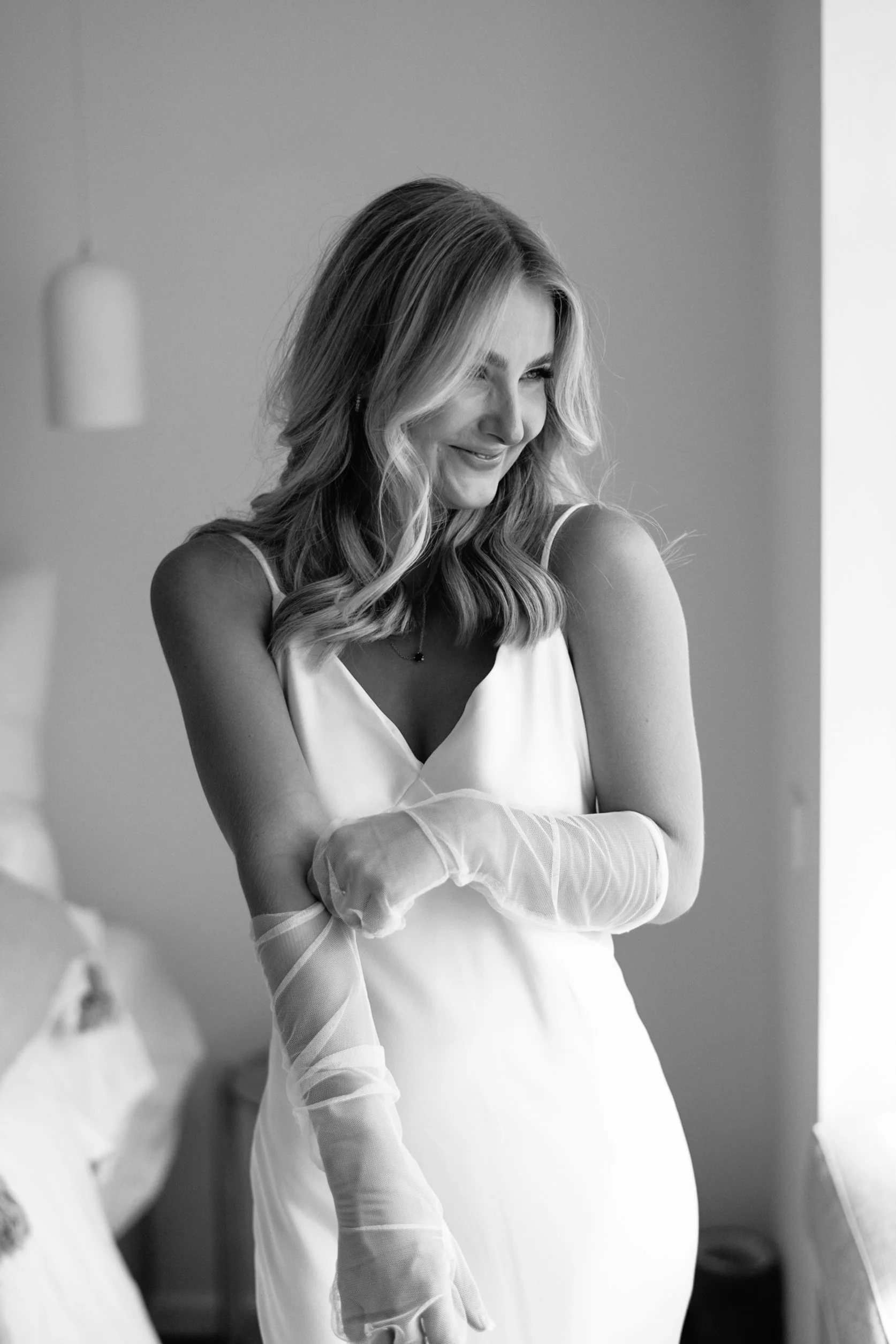 A smiling woman with wavy, shoulder-length hair wearing a sleeveless white dress and sheer gloves, standing indoors against a plain wall.