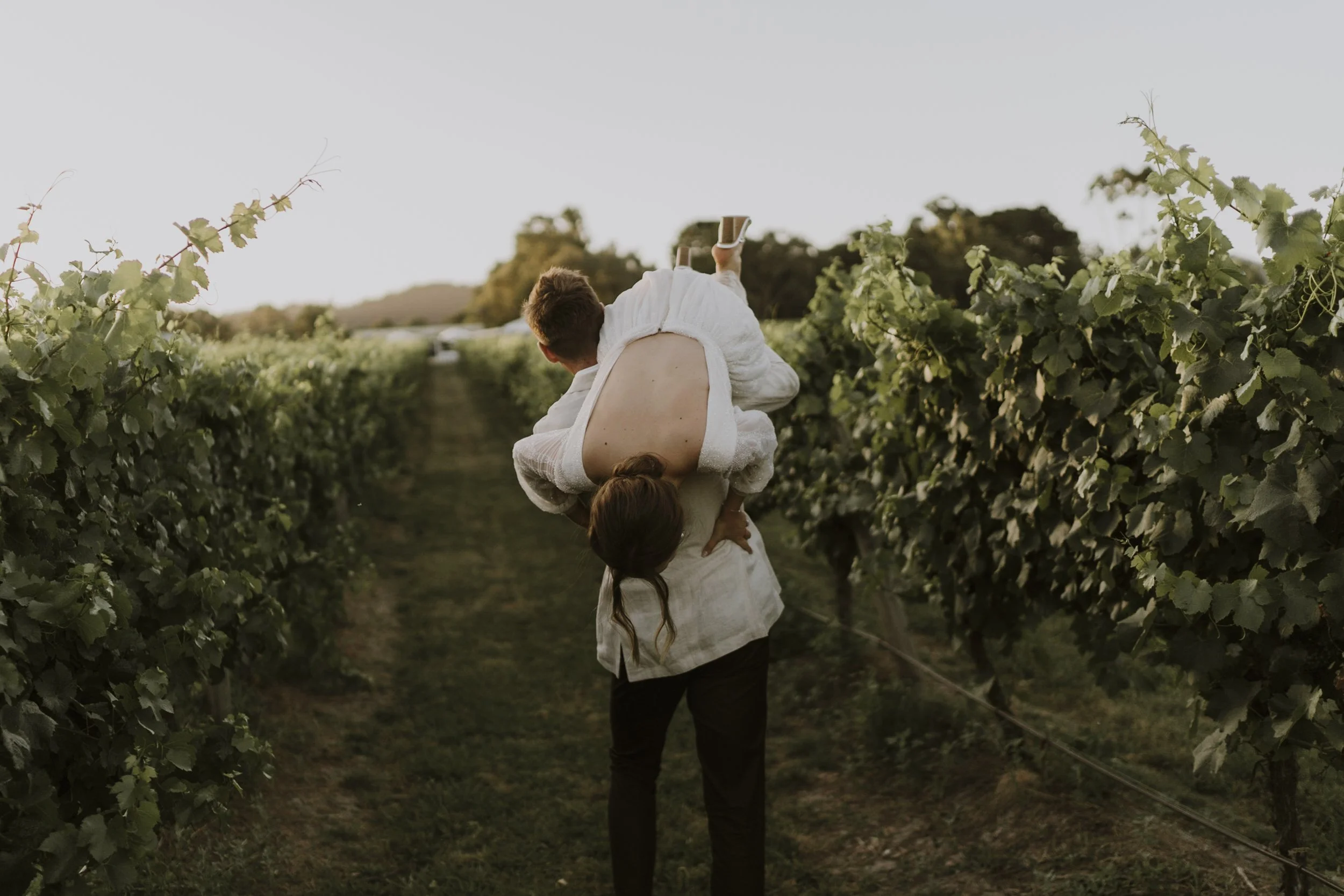 A person holding a child upside down in a vineyard during sunset, both wearing white clothing.