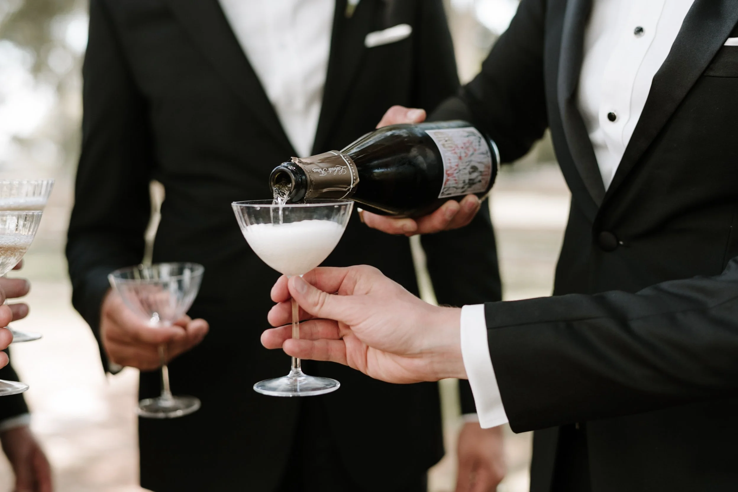 People in tuxedos are at an outdoor event, with one person pouring champagne into a glass.