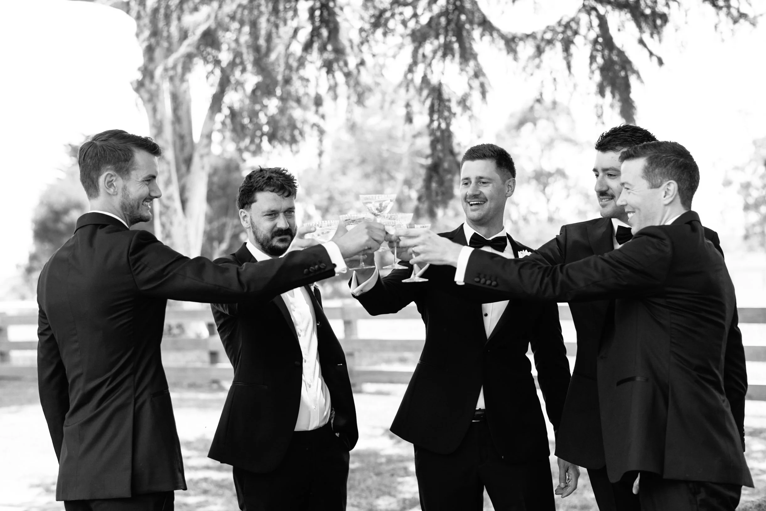 Six men dressed in tuxedos raising glasses in a toast outdoors, with trees in the background.