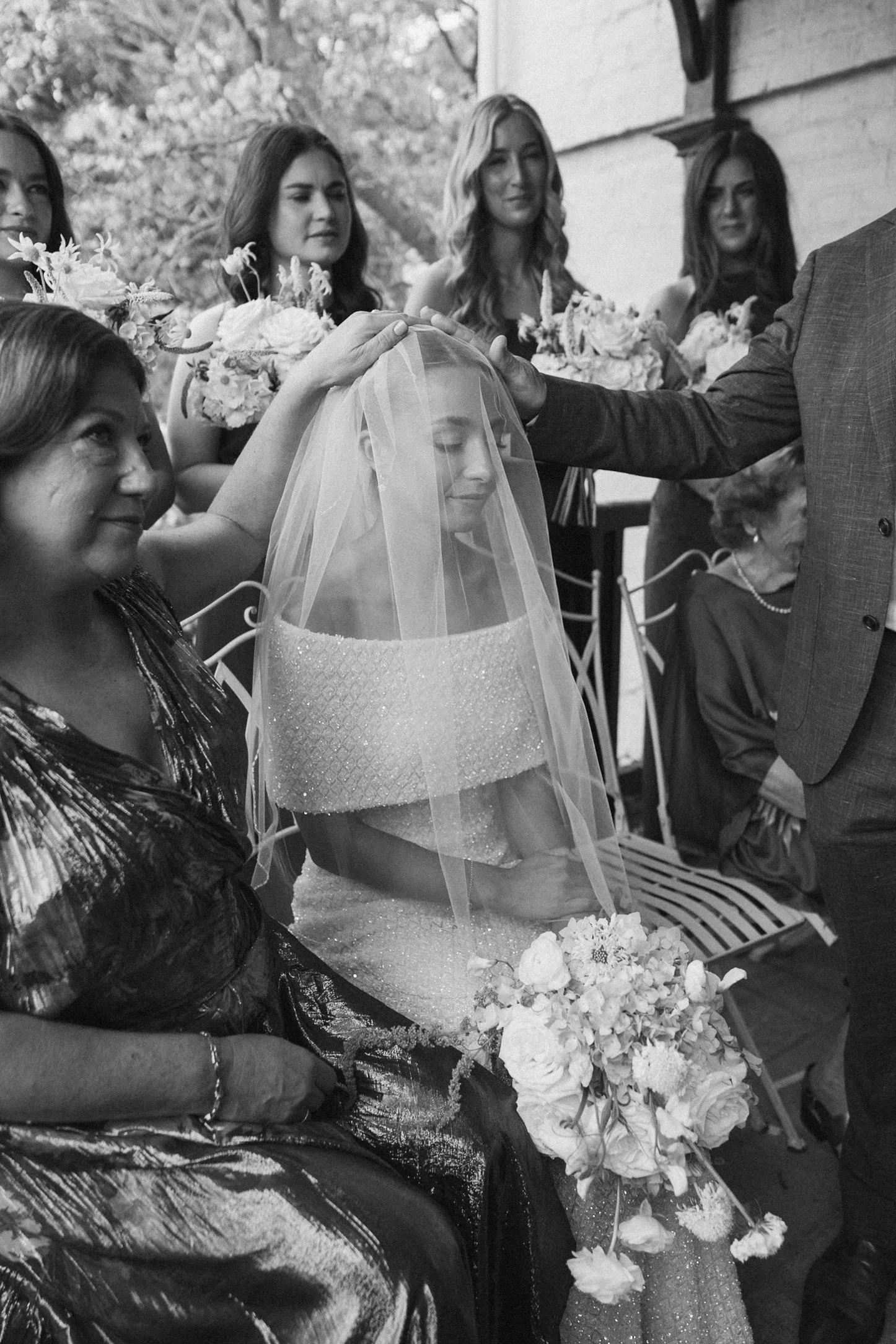 A black and white photo showing a bridal party at a wedding. The bride, wearing a veil, is seated with her eyes closed. Several women, likely bridesmaids, are standing behind her, holding bouquets of flowers. A man in a suit is gently resting his han