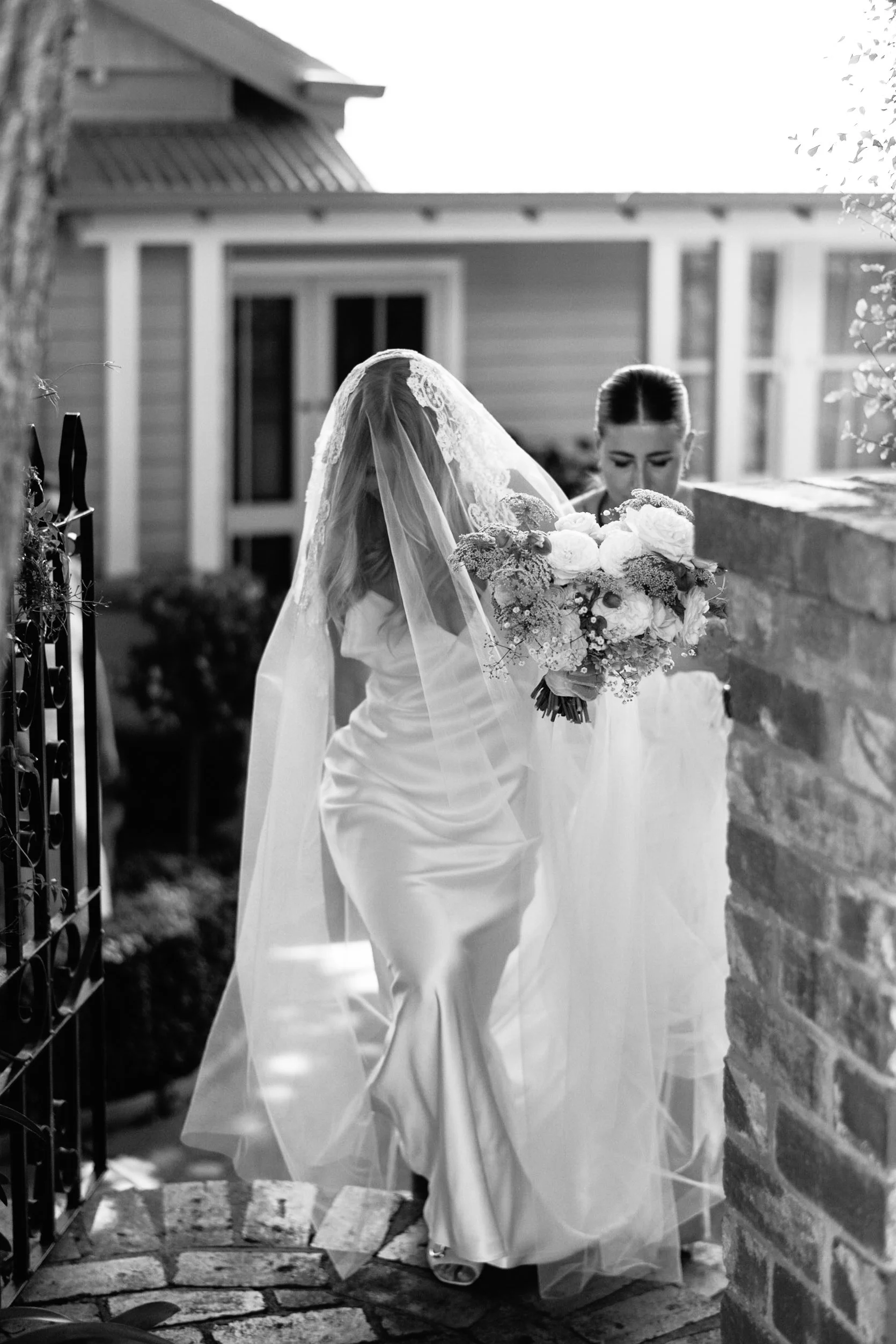 A bride in a white wedding dress with a veil and a woman holding a bouquet of flowers standing outside near a brick wall, with a house in the background. The scene is black and white.