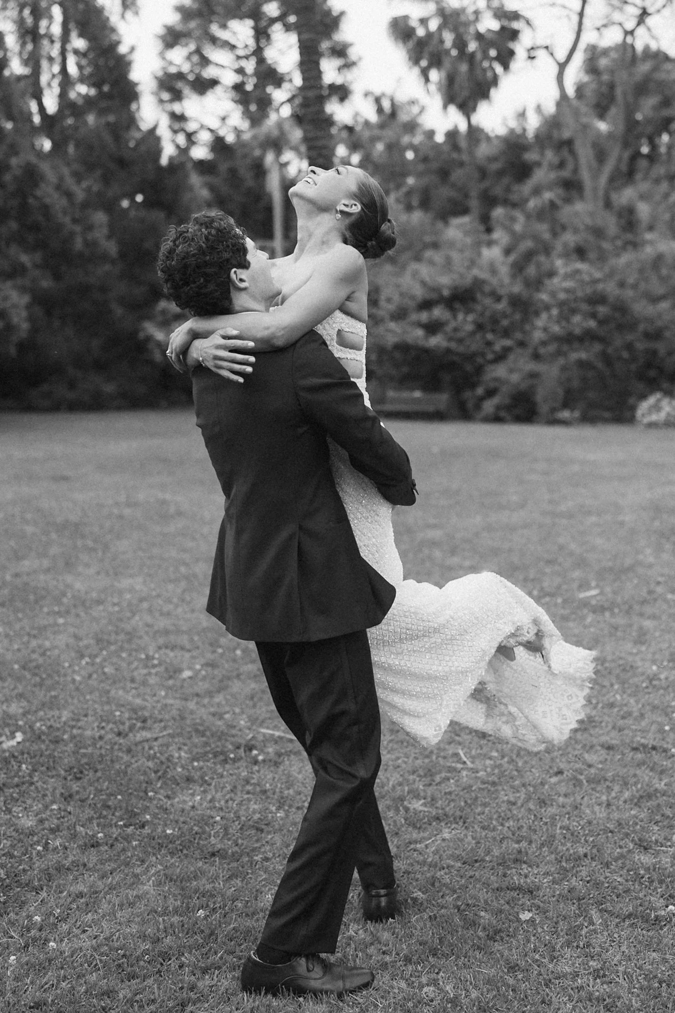 A man in a suit lifting a woman in a wedding dress in his arms outdoors, with trees in the background, on a grassy field, captured in black and white.