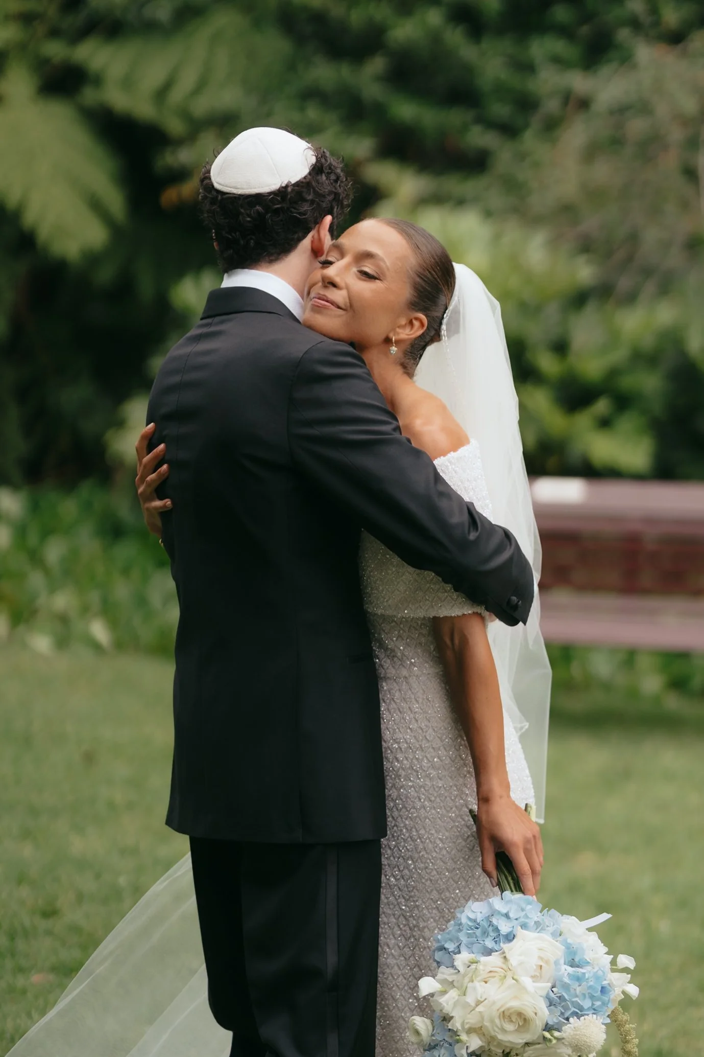 A bride and groom hugging outdoors, the bride holding a bouquet of white and blue flowers, with a veil and wedding dress, and the groom wearing a black suit and kippah.