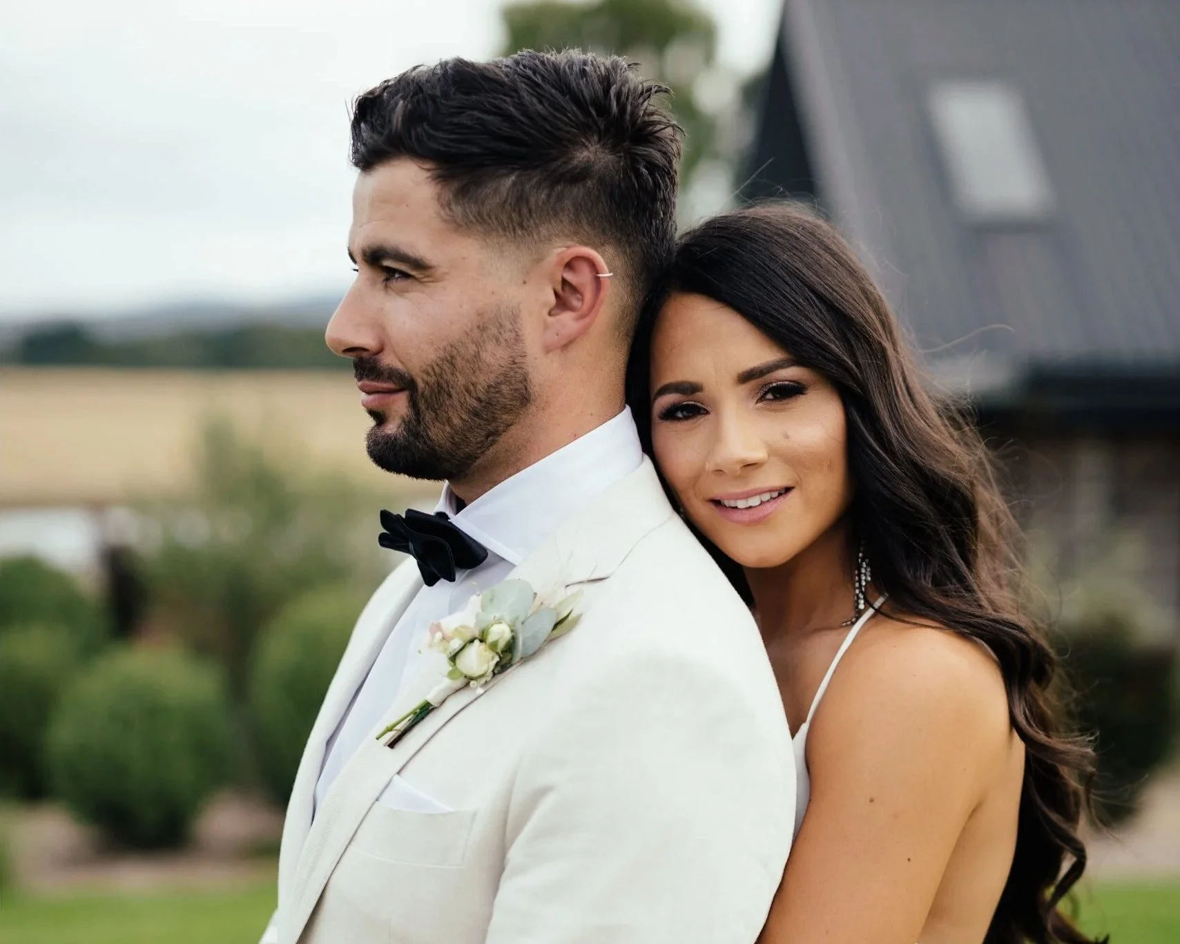 A bride with dark hair and a groom with a beard are standing outdoors. The bride is smiling and leaning her head on the groom's shoulder, who is facing sideways. The groom is wearing a white tuxedo with a black bow tie and a boutonniere with white flowers. The background is blurred with greenery and a dark structure.