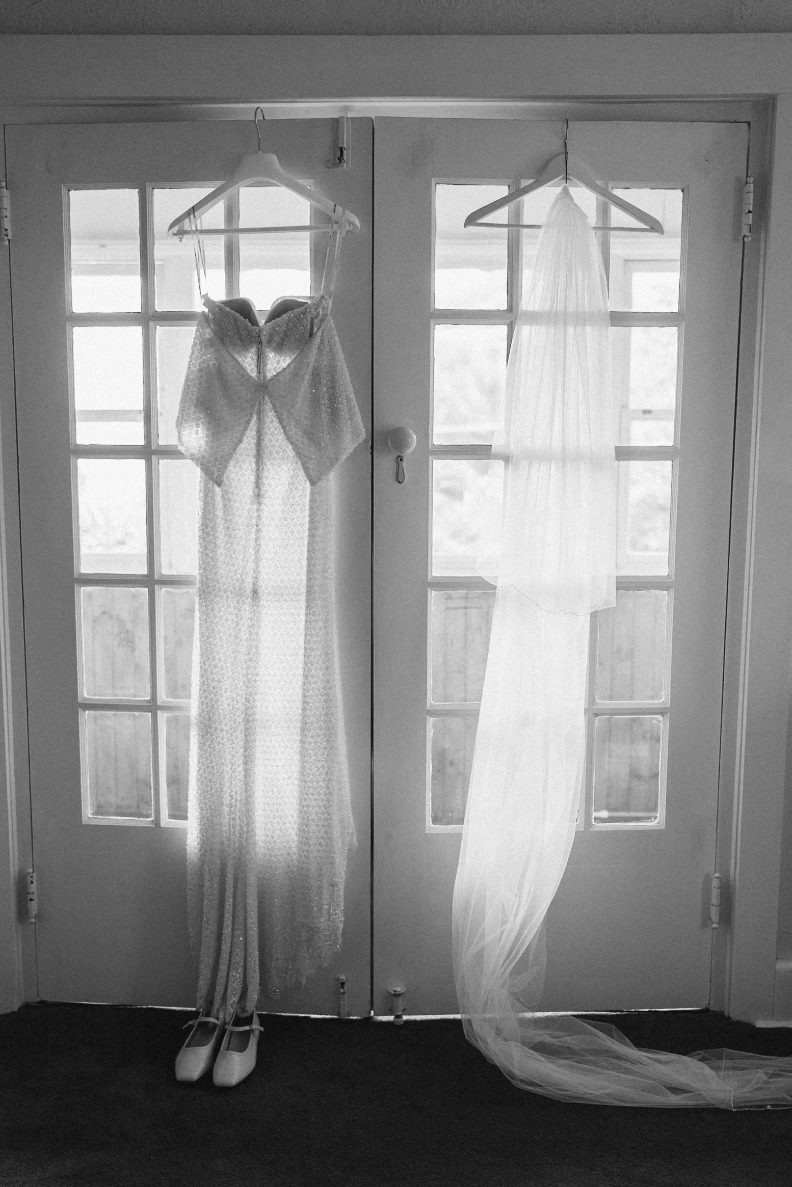 Two wedding dresses hanging on white hangers on a glass-paneled door, with white shoes and a veil on the floor in front of the dresses.