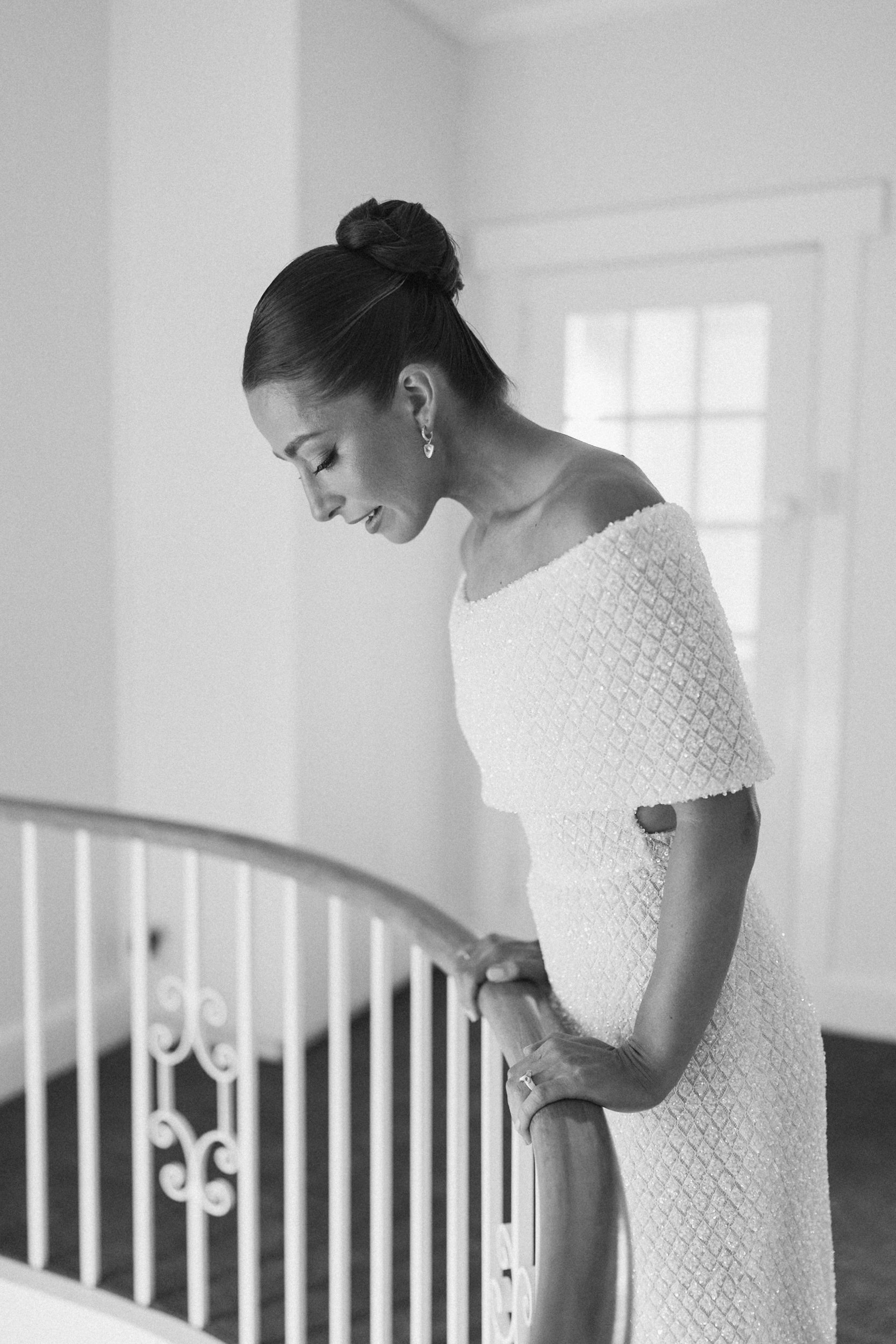 Black and white photo of a woman in an elegant dress, with her head bowed, standing beside a white decorative banister.