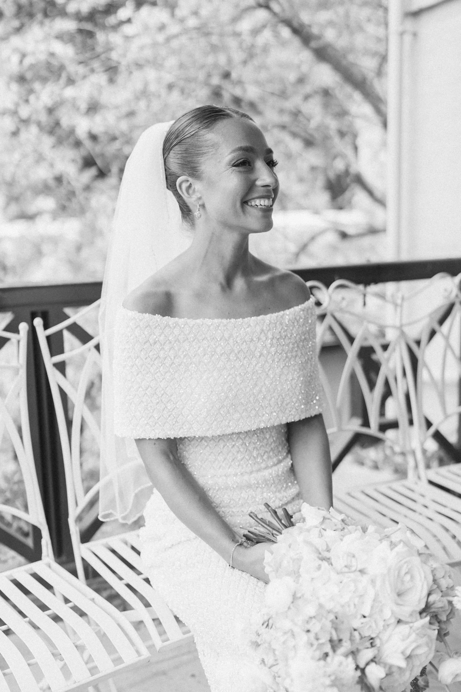 Black and white photo of a smiling woman in an off-the-shoulder dress holding a bouquet of flowers, sitting outdoors near white chairs, with trees in the background.