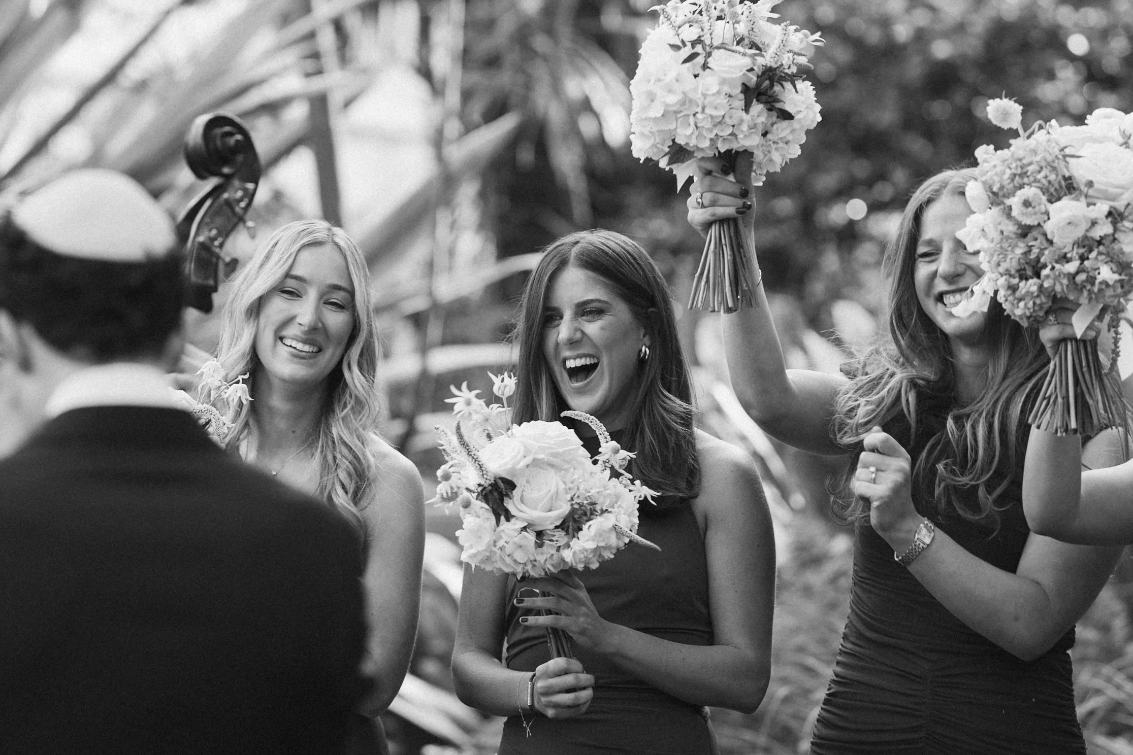 A group of women celebrating outdoors, some holding bouquets of flowers, smiling and appearing joyful, with a man in the foreground facing them.