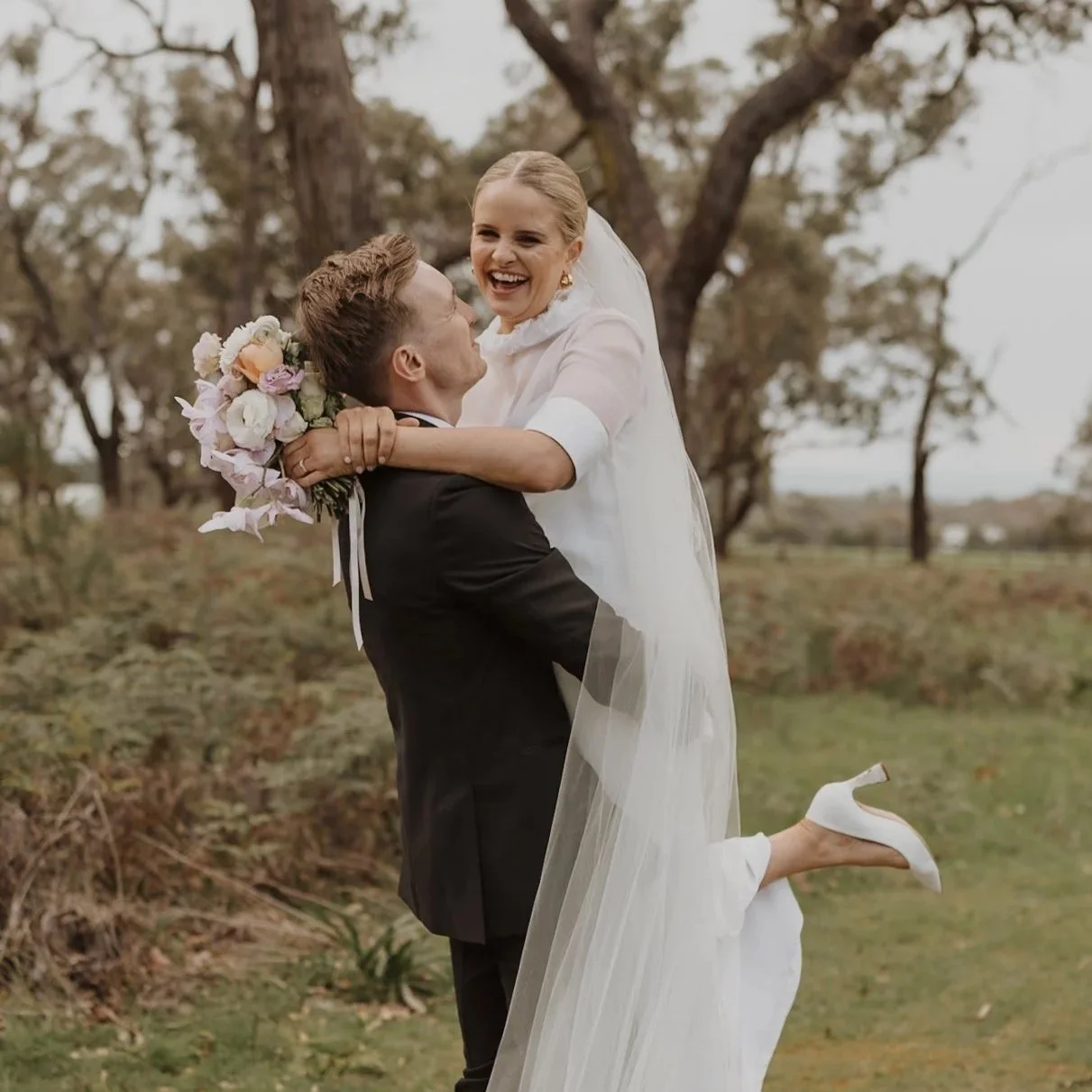 A bride and groom celebrating outdoors, with the groom lifting the bride. The bride is smiling, holding a bouquet of flowers, and wearing a white wedding dress. The groom is wearing a black suit. The background features trees and a natural landscape.