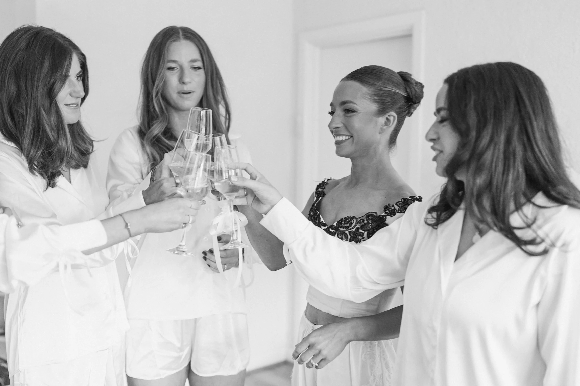 Women toasting with champagne glasses at a celebration event, some women wearing white outfits, one woman with an ornate top, smiling and enjoying the moment.