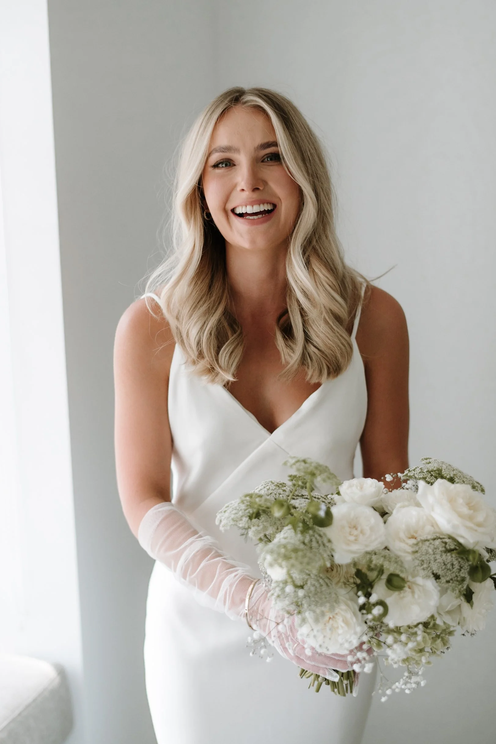 A smiling woman with blonde wavy hair wearing a white dress and gloves holding a bouquet of white flowers.