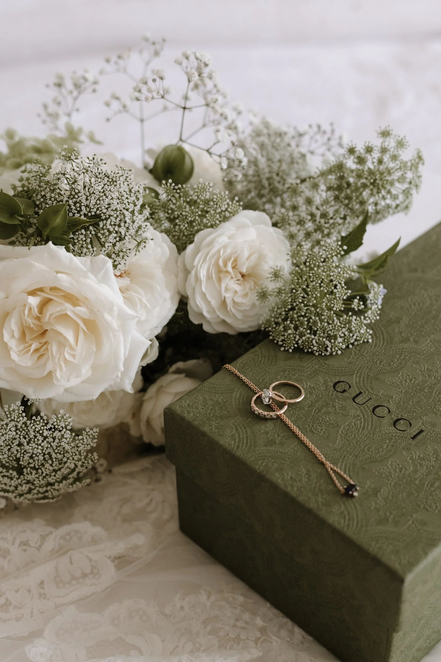 Cream-colored flowers and greenery next to a green Gucci jewelry box with a ring and a necklace on top.