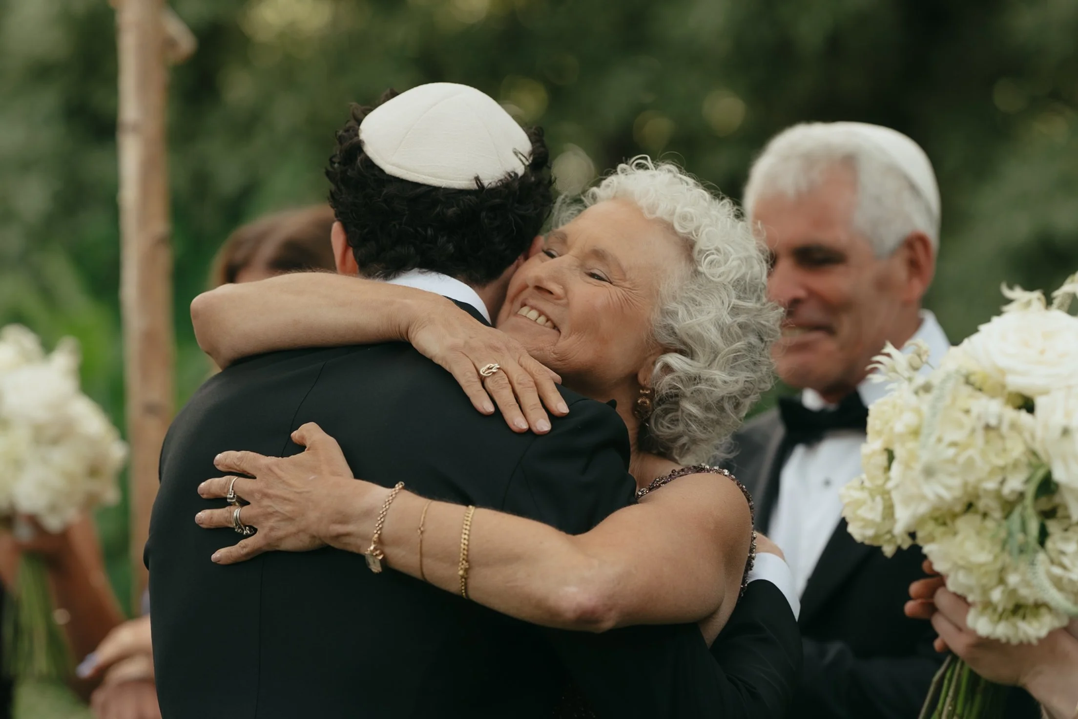 A woman with curly gray hair hugging a man wearing a dark suit and a white yarmulke at an outdoor wedding ceremony, with a man in a tuxedo and bow tie visible in the background holding white flowers.