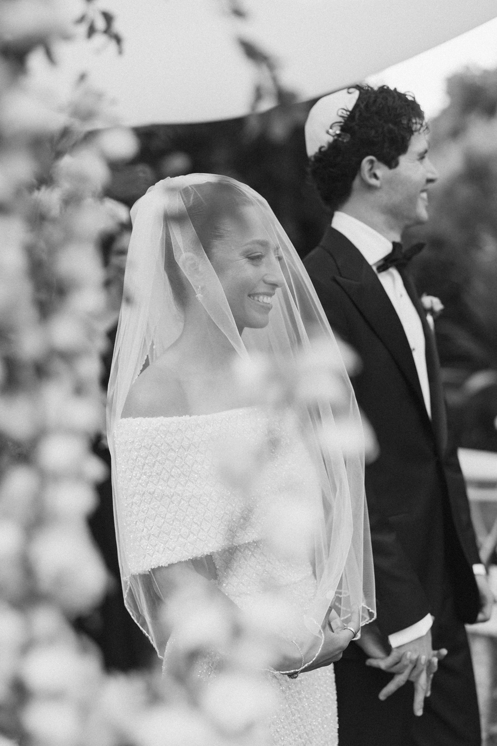 Black and white photo of a smiling bride in a wedding dress with a veil and a groom in a tuxedo holding hands during a wedding ceremony.