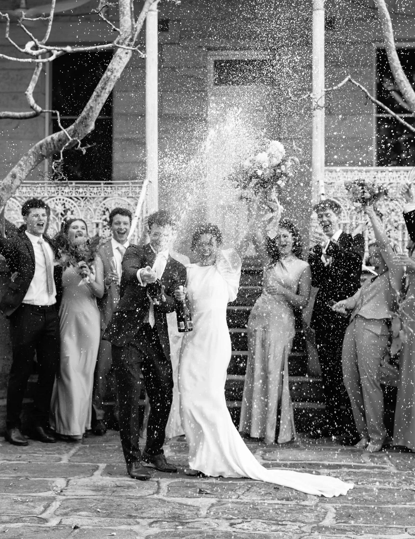 A bride and groom celebrating their wedding with friends and family, splashing champagne in the air outdoors, during daytime.