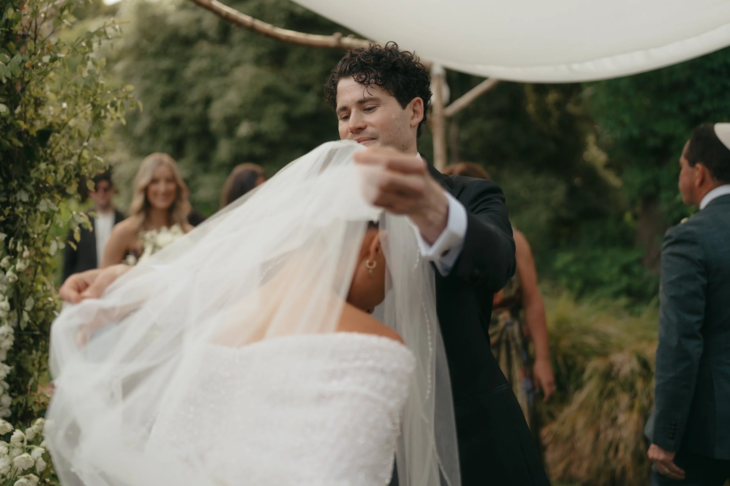 A man in a black suit holding the train of a wedding dress at an outdoor wedding ceremony.