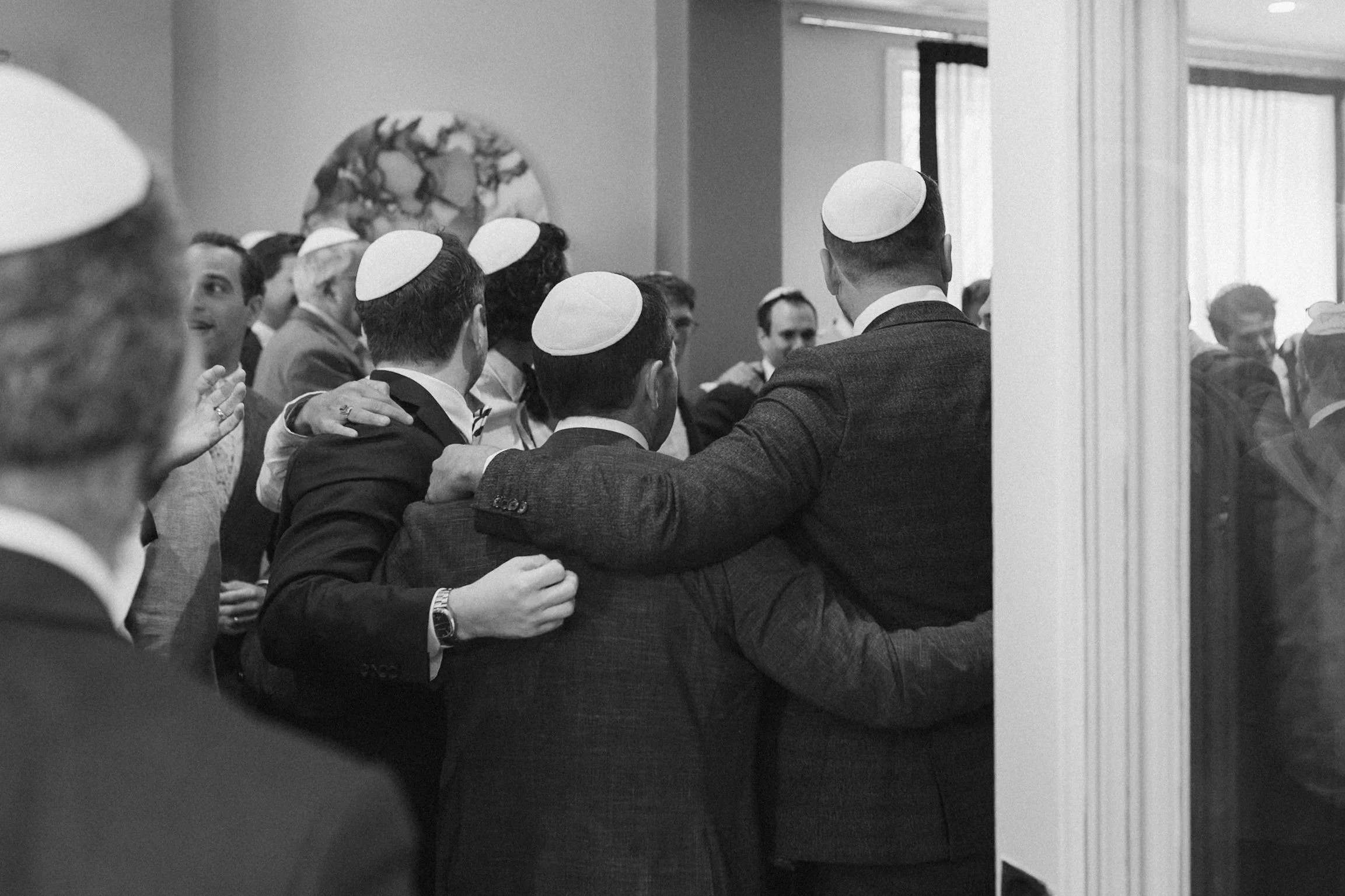 A group of men wearing suits and kippahs (Jewish skullcaps) engaging in a group prayer or religious activity in a room, with a sculpture or artwork on the wall in the background.