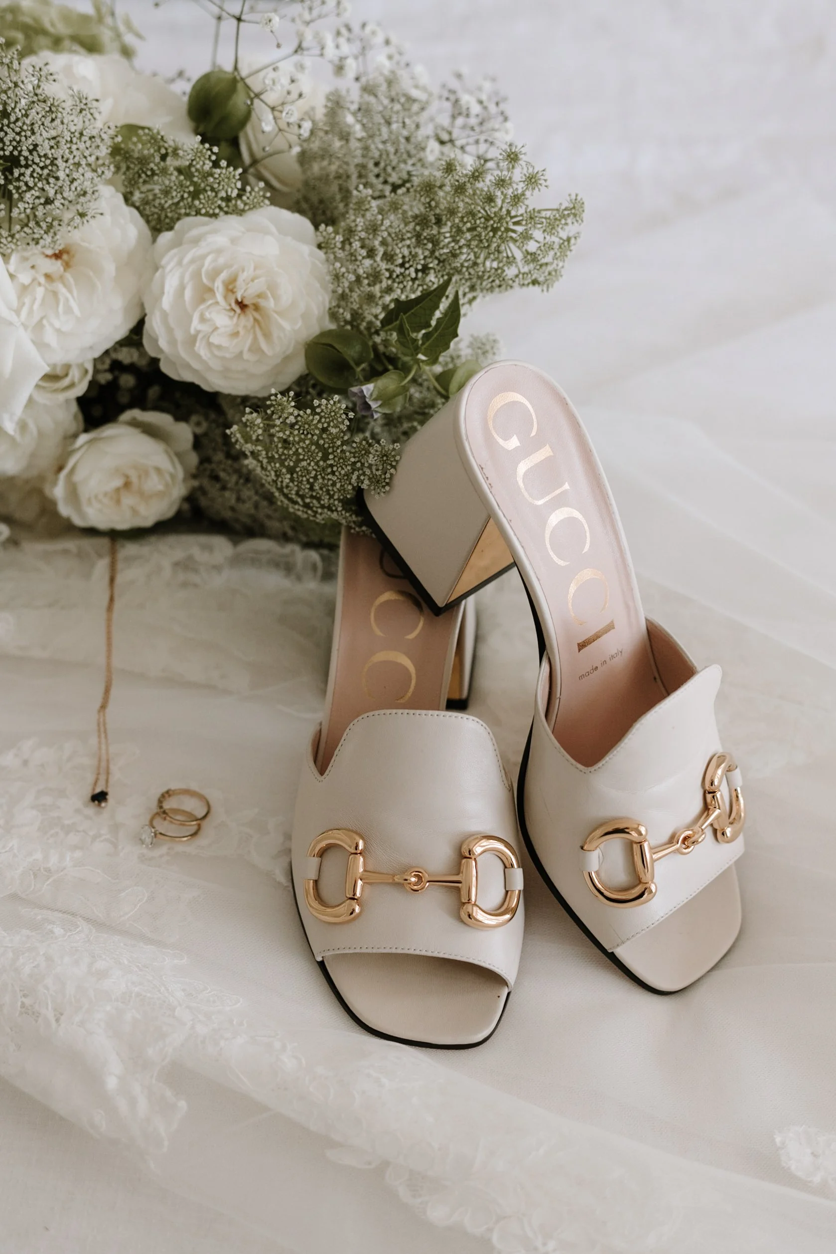 A pair of white Gucci high-heeled shoes with gold hardware, a bouquet of white flowers and greenery, a lace fabric, a gold ring, and a gold bracelet on a white surface.