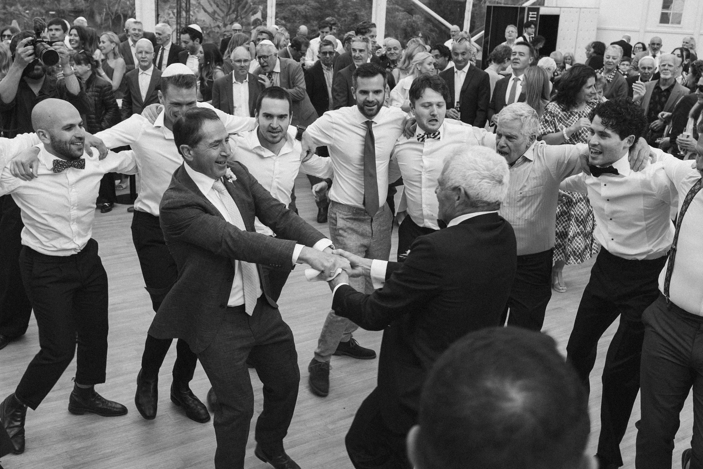 A group of men and women dancing in a circle at a celebration, with some wearing yarmulkes and formal attire, surrounded by a crowd of onlookers.