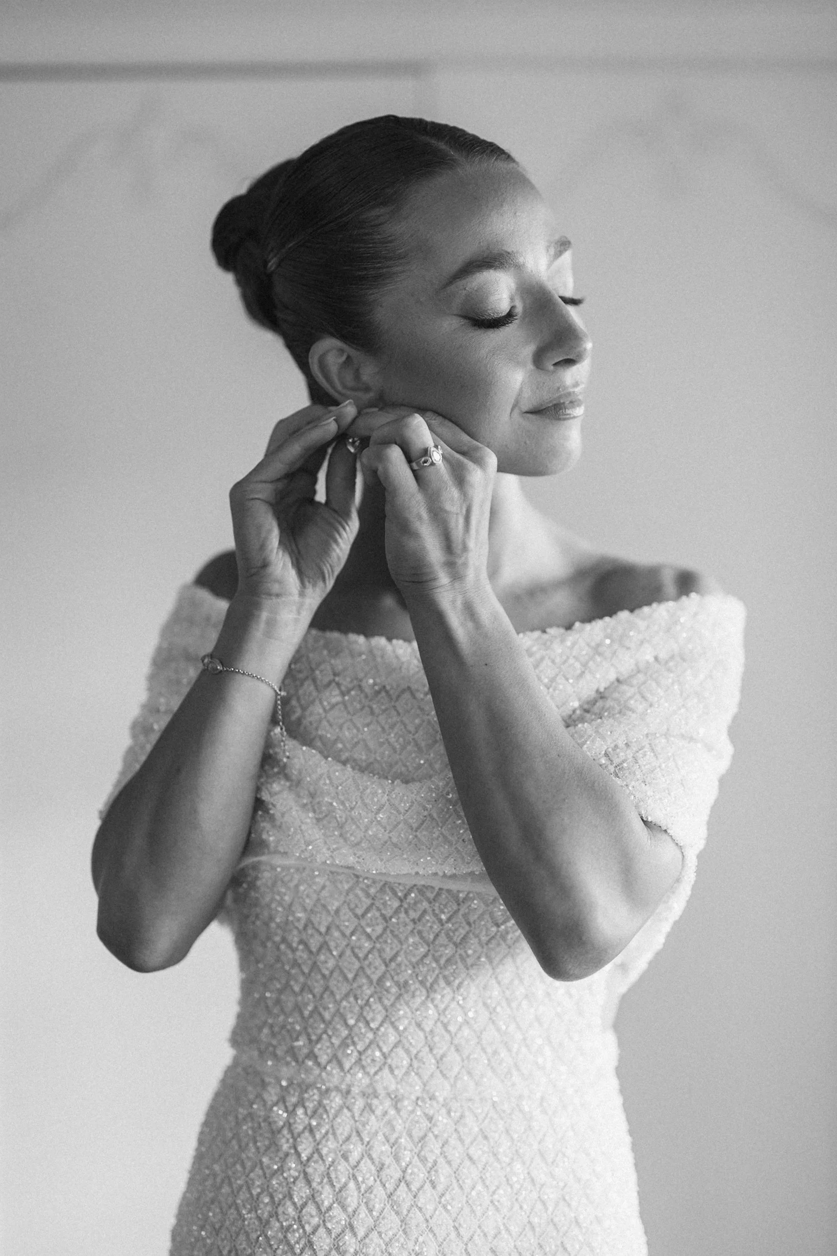 A woman in a textured dress with her eyes closed, adjusting an earring, wearing jewelry, with her hair styled in an elegant updo.
