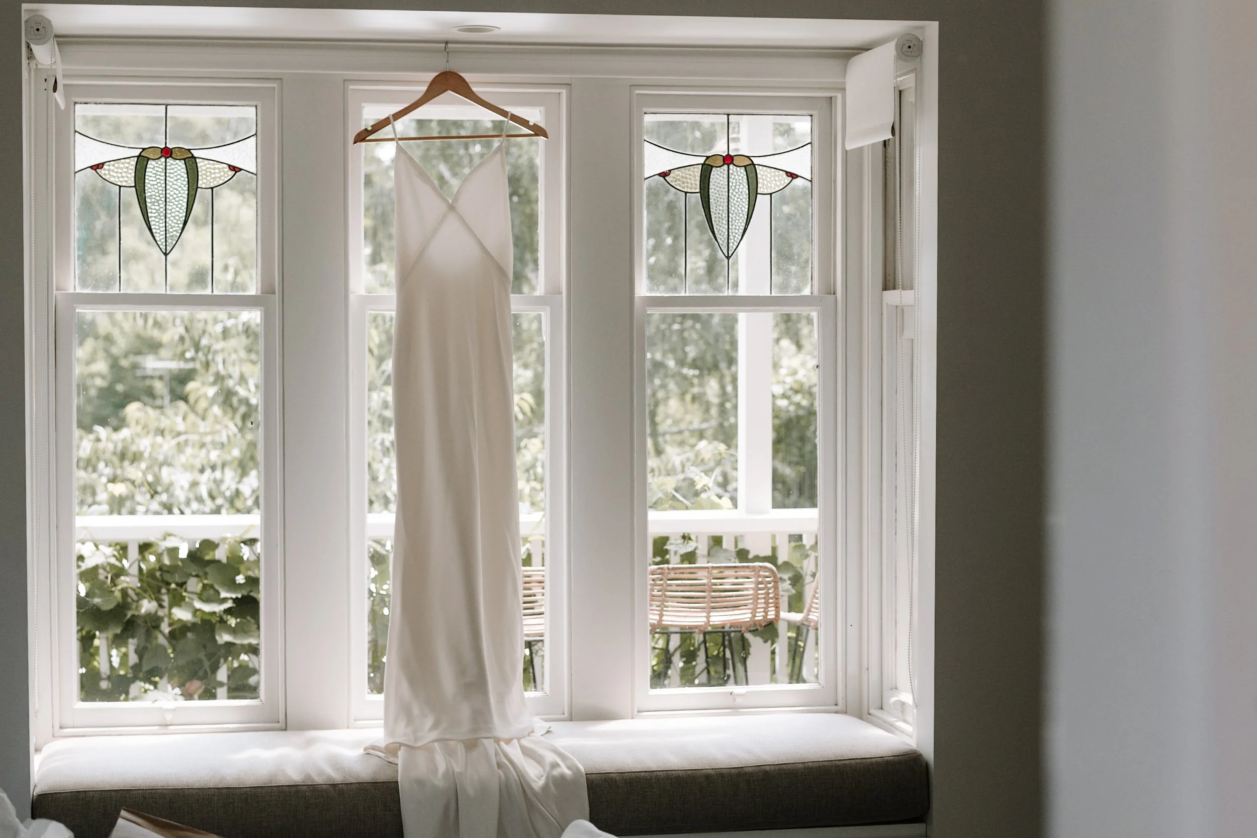 A white wedding dress hanging on a wooden hanger in front of a large window with stained glass accents. Outside, there are green trees and a wooden bench.