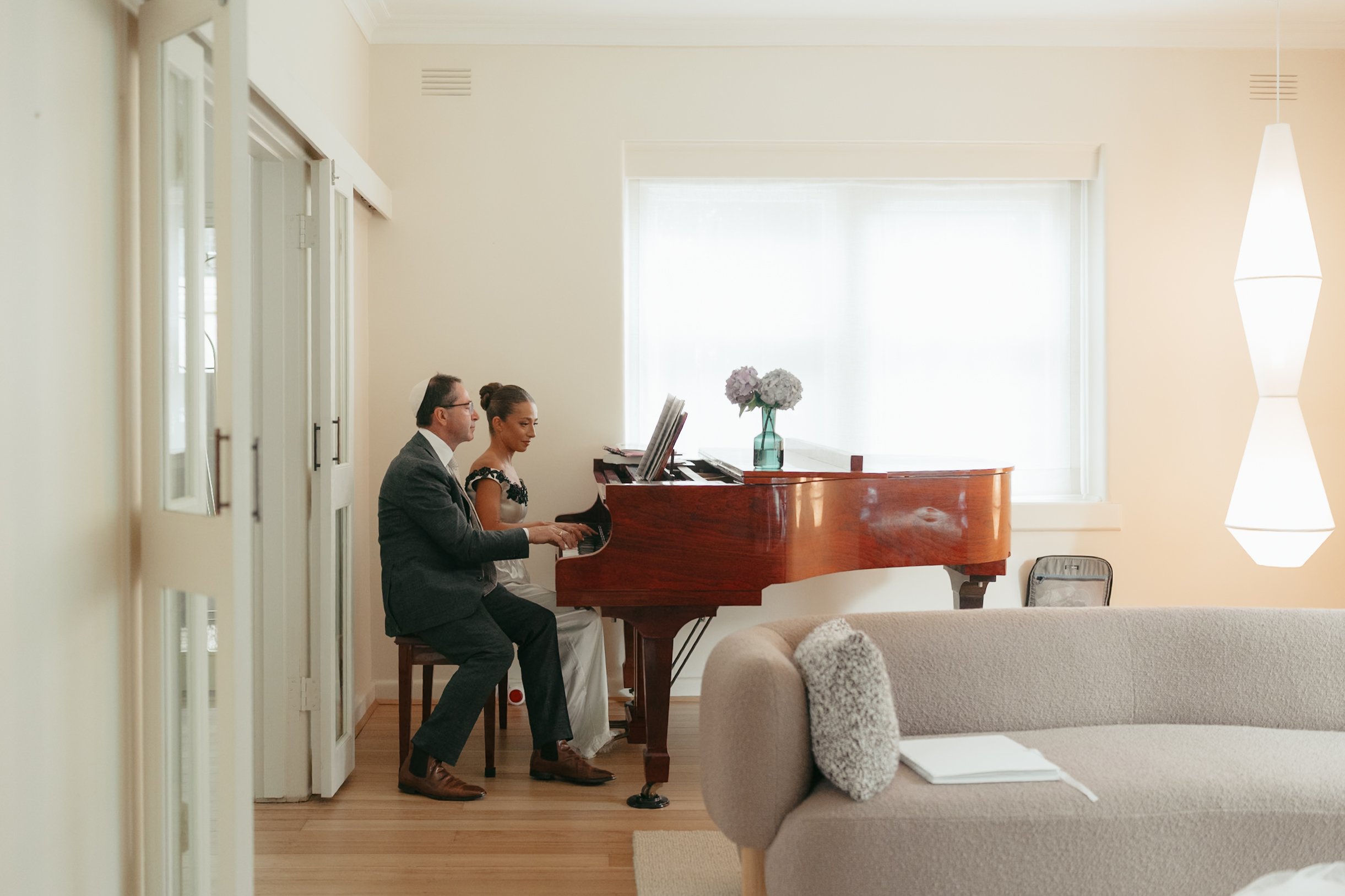 A man and woman playing a grand piano in a bright, airy living room with a large window, a vase with flowers on the piano, and a modern hanging light fixture.