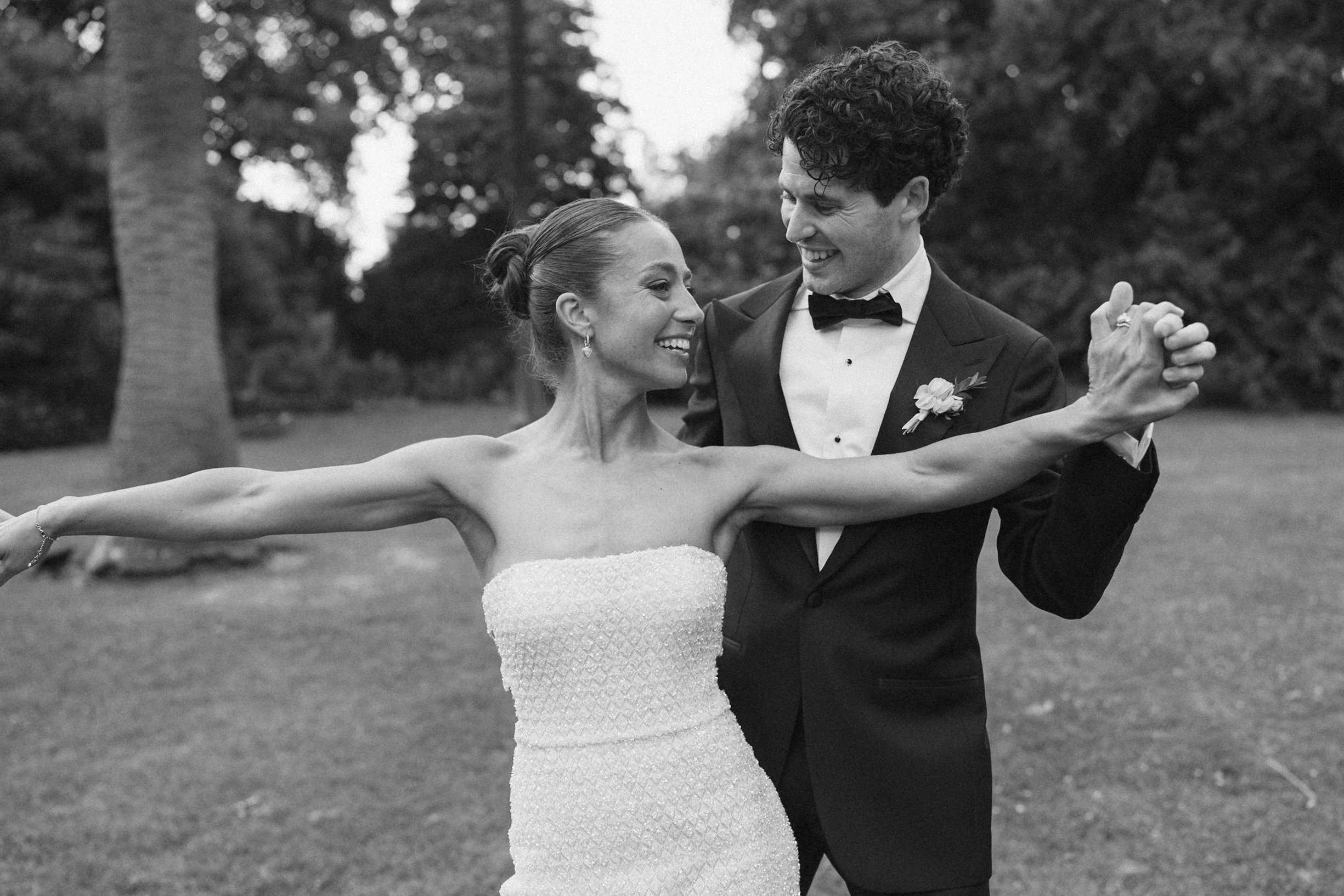 A black and white photo of a bride and groom dancing outdoors, smiling and looking at each other, with trees in the background.