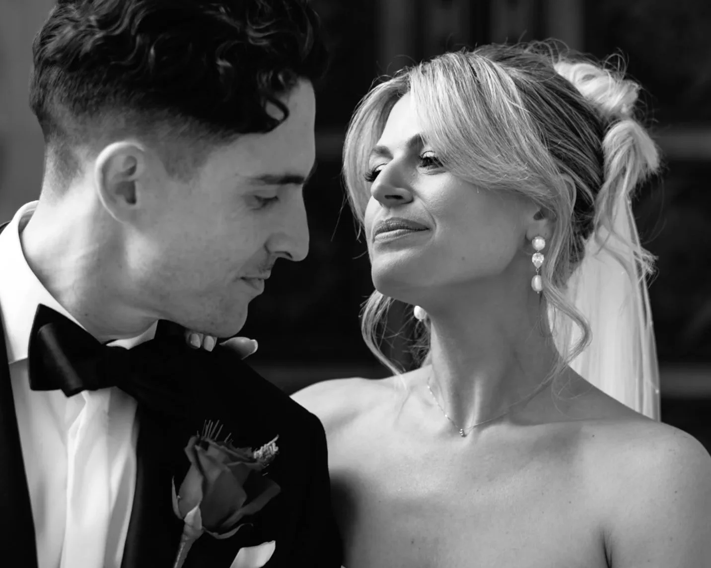 Black and white photo of a man and woman closely facing each other, possibly at a wedding, with the man in a tuxedo and the woman with earrings and wavy hair.