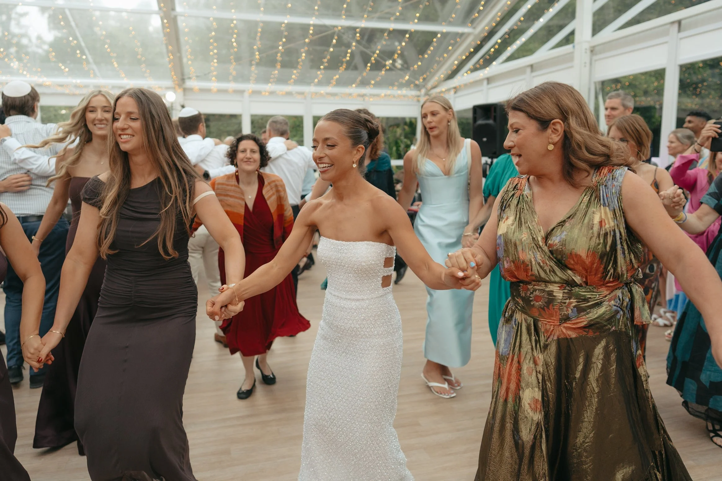 People dancing and celebrating at a wedding reception under a glass pavilion with fairy lights, featuring women in formal dresses and a bride in a white gown.