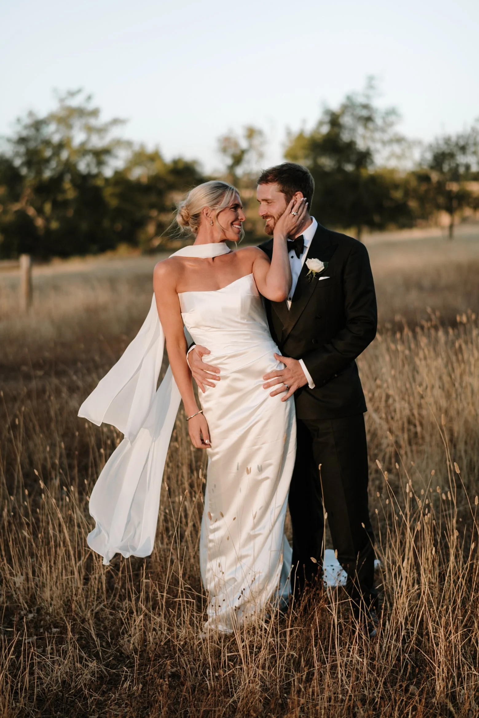 A bride and groom in formal wedding attire standing in a field at sunset, gazing into each other's eyes and smiling.