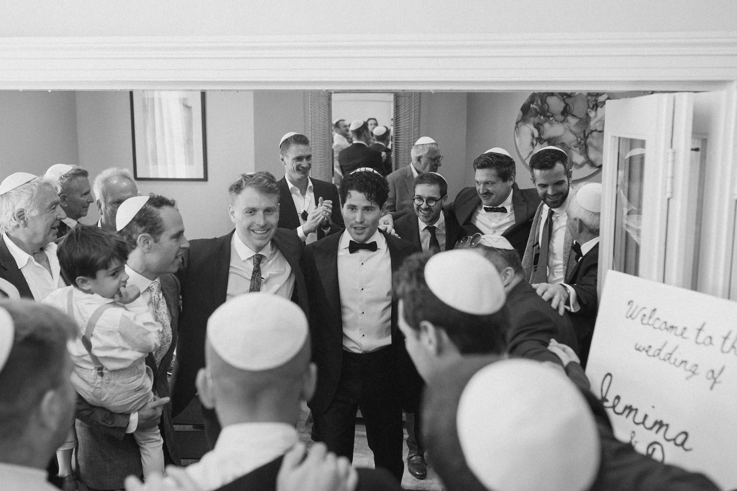 Group of men and boys attending wedding reception, wearing yarmulkes, dressed in suits and bow ties, gathered in a room with artwork and a sign that reads "Welcome to the wedding," smiling and socializing.