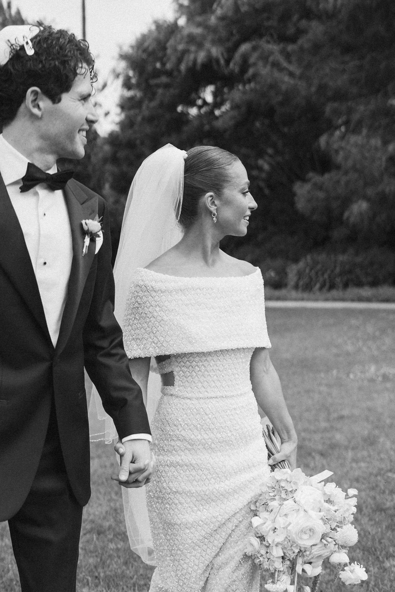 Black and white photo of a newlywed couple holding hands outdoors, smiling and walking, with trees in the background. The groom wears a tuxedo with a bow tie and boutonniere, and the bride wears a textured wedding gown, a veil, and drop earrings, hol